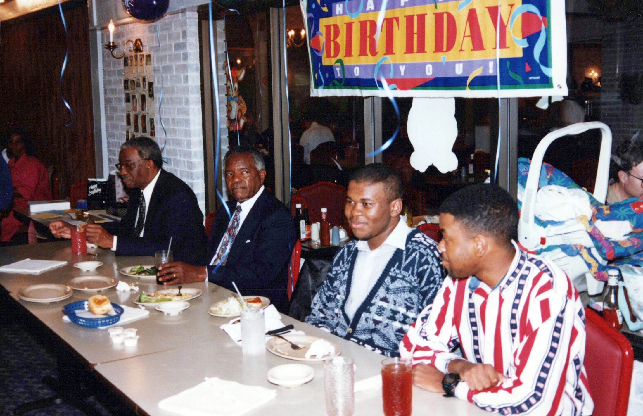 Four Black men, two older in suits and two younger in sweaters, sit at a long table in a restaurant with a colorful "Happy Birthday" banner hanging above them.