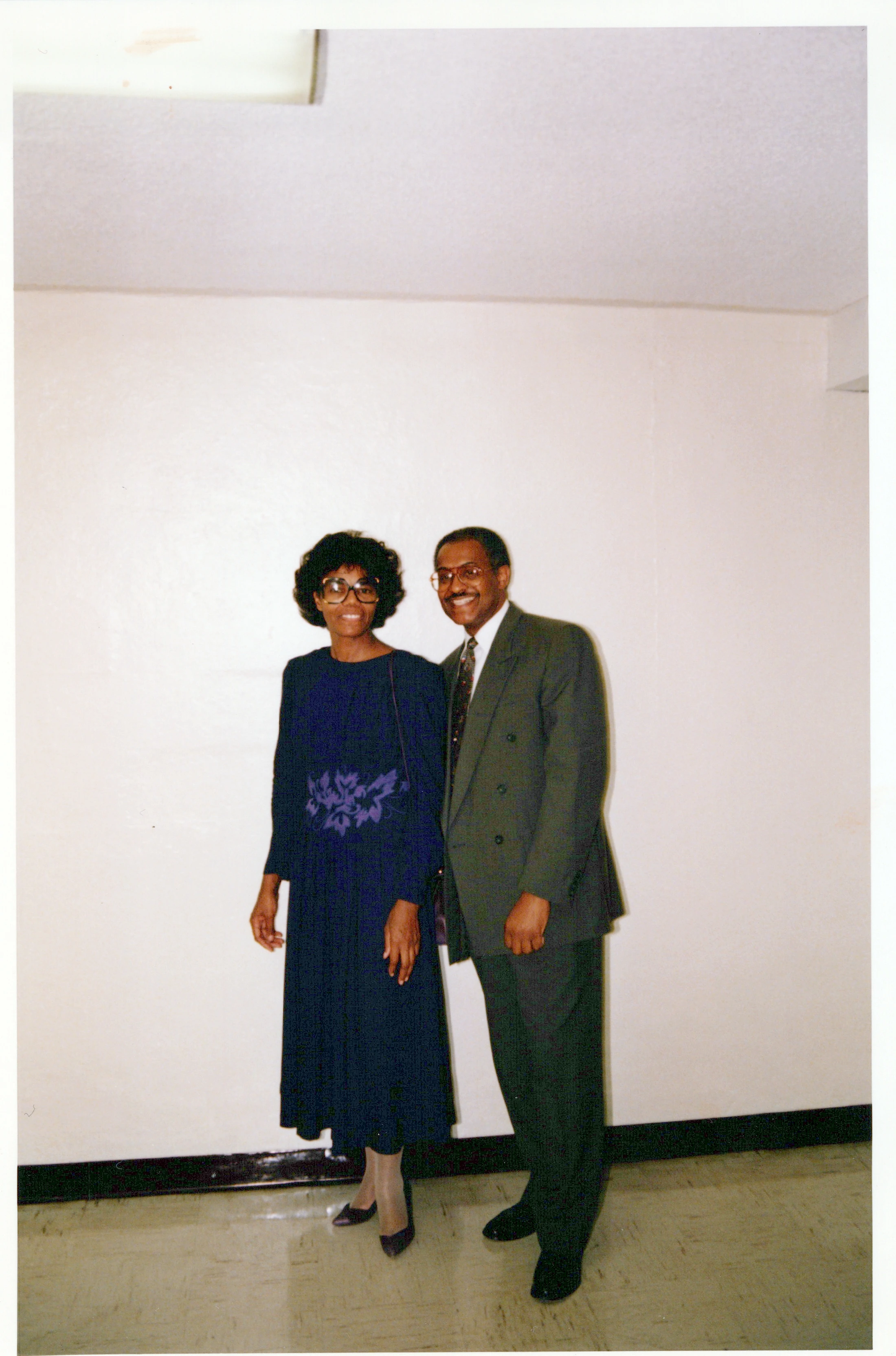 A vintage, full-length photograph of a smiling African American couple. The woman is in a dark blue dress, and the man is in a dark green suit and tie.