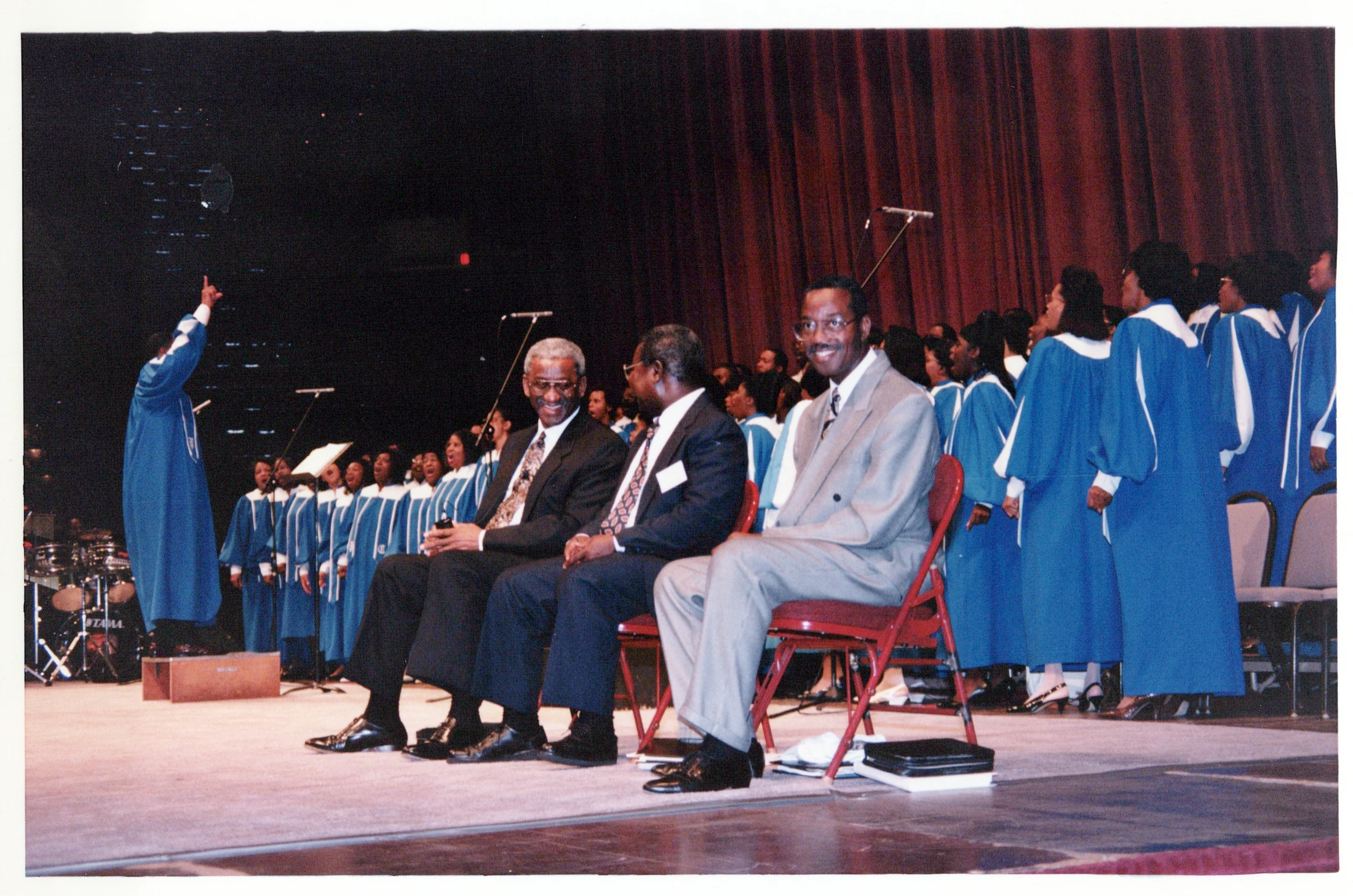 Three men in suits sit in the front row on a stage, watching a large gospel choir in blue and white robes perform behind them.
