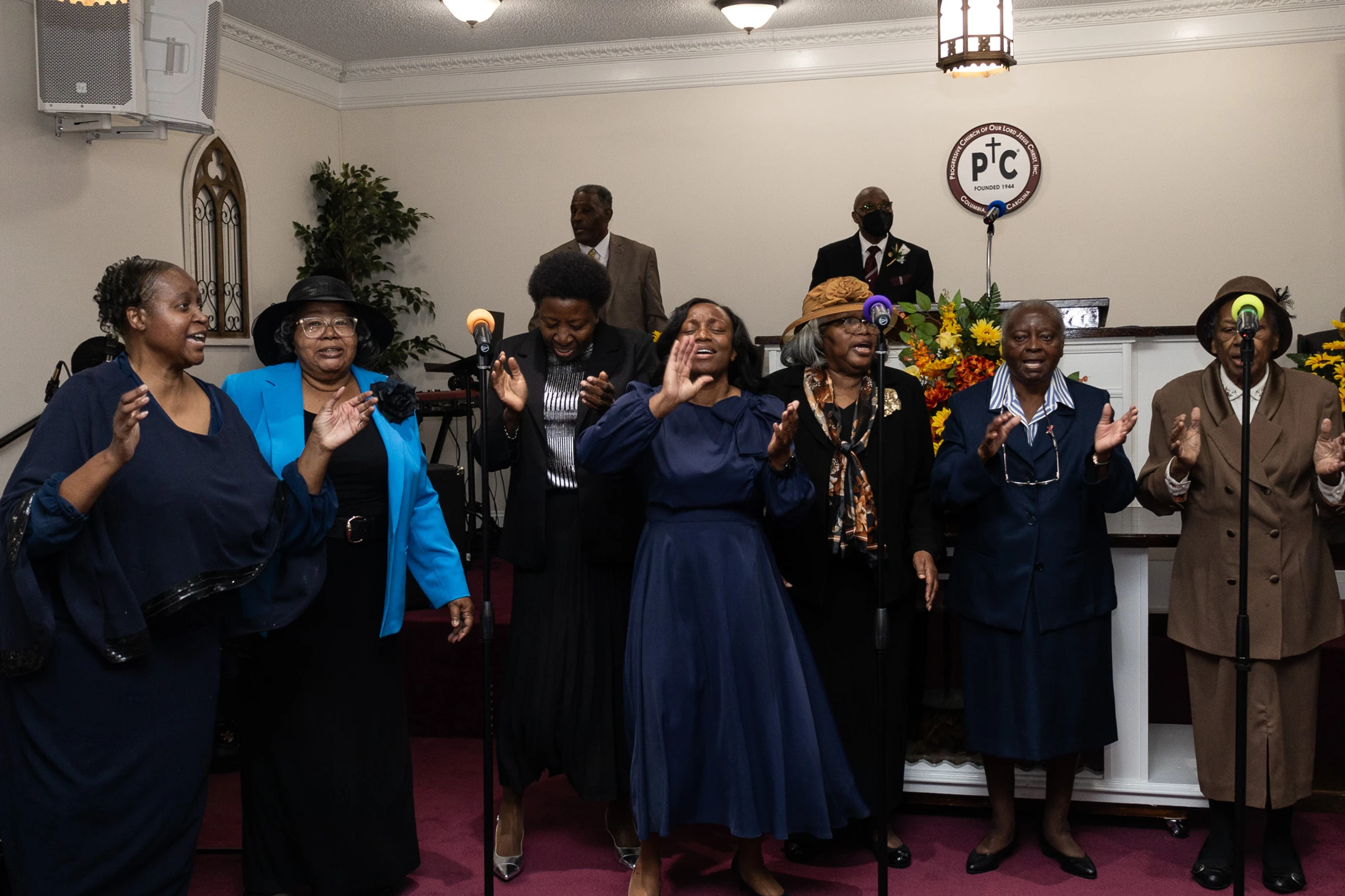 A group of Black women in a church choir joyfully sing and clap their hands, standing in front of microphones during a worship service.