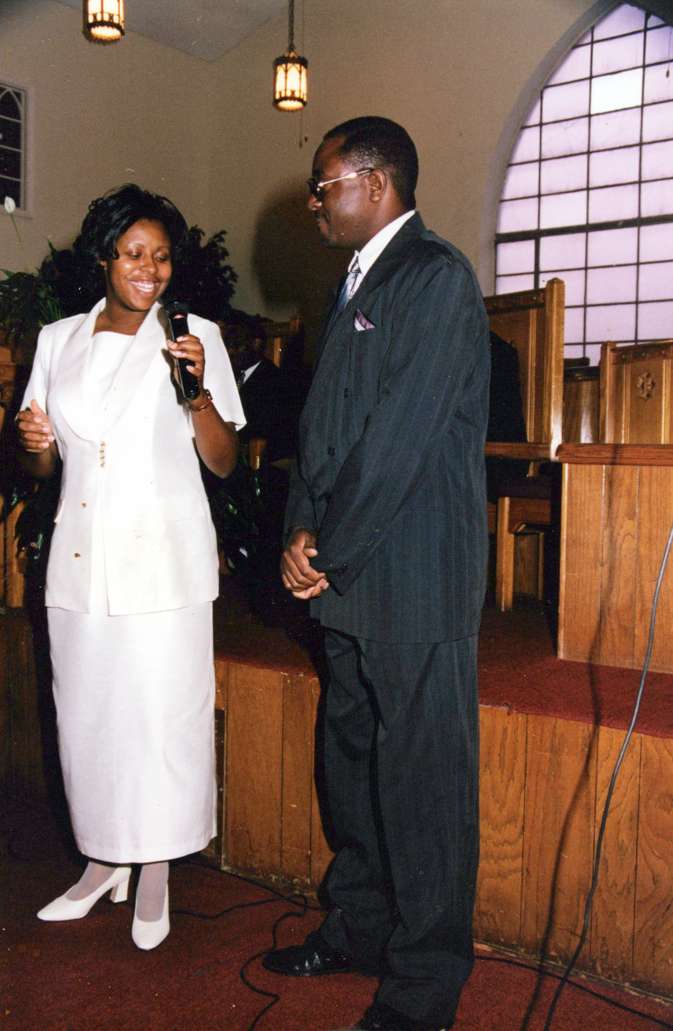 A vintage photo shows a Black woman in a white suit smiling while holding a microphone, standing next to a Black man in a dark pinstripe suit inside a church.