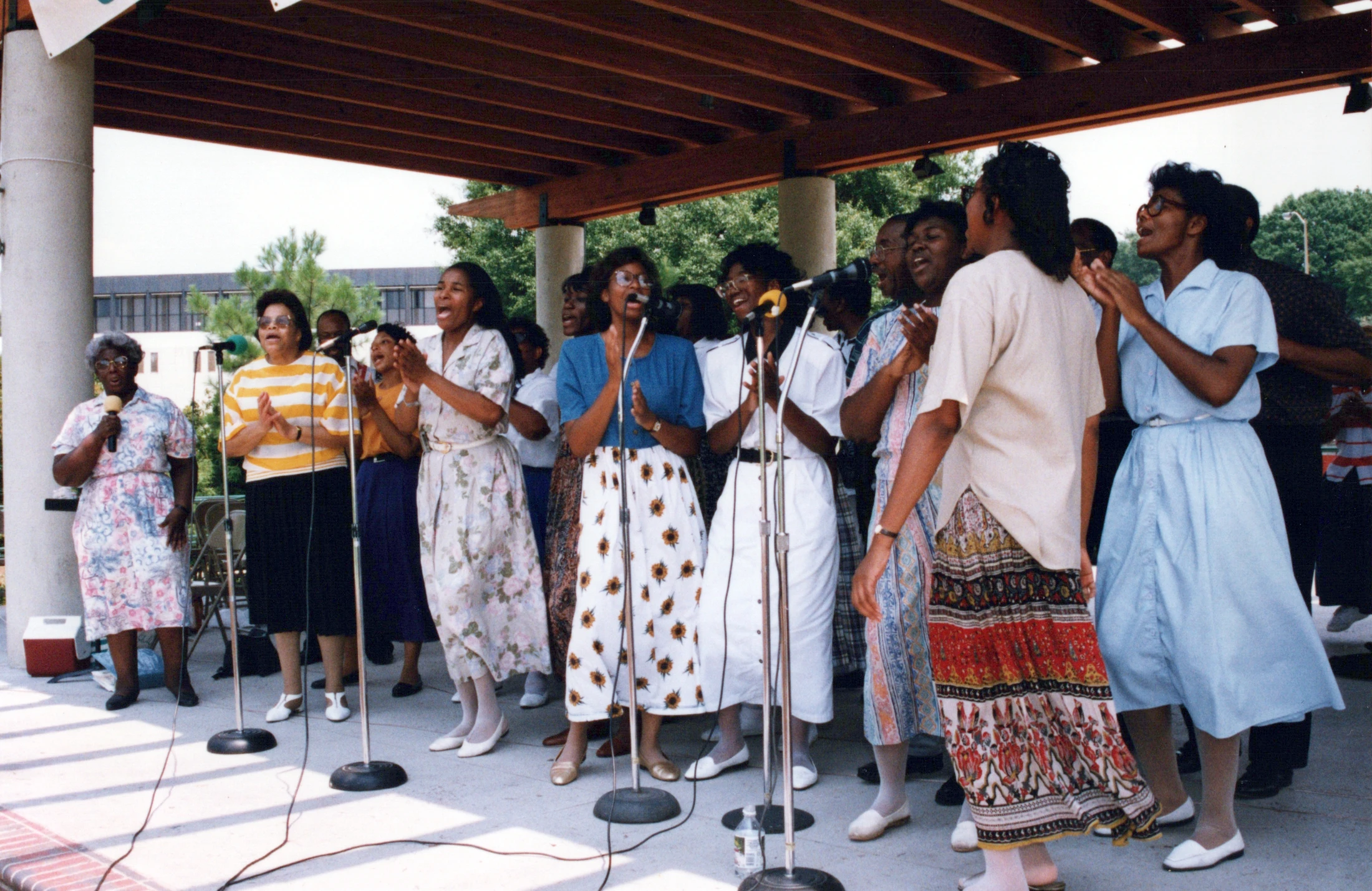 A group of Black women in dresses and skirts sing enthusiastically into microphones on an outdoor stage, clapping their hands.