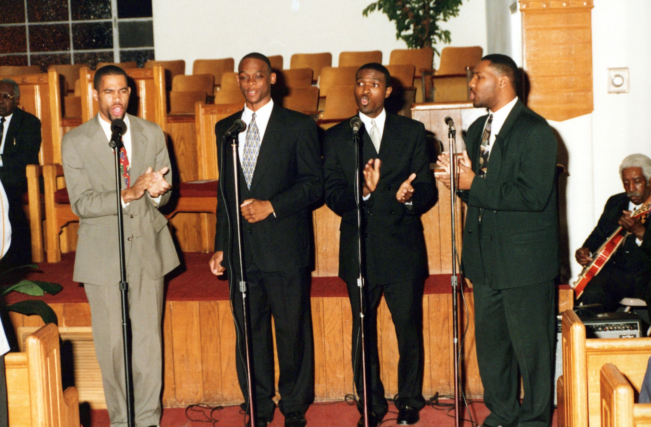 Four Black men in suits stand in a line, singing into microphones and clapping during a performance in a church. A guitarist plays in the background.