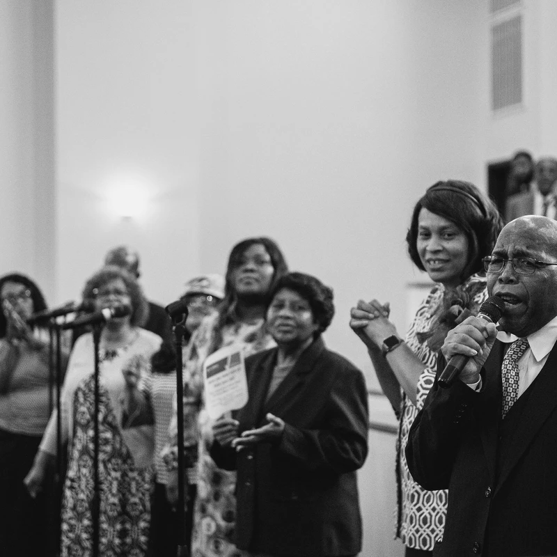 A black and white photo of an older Black man in a suit singing into a microphone, with a choir of smiling women standing and singing behind him in a church.