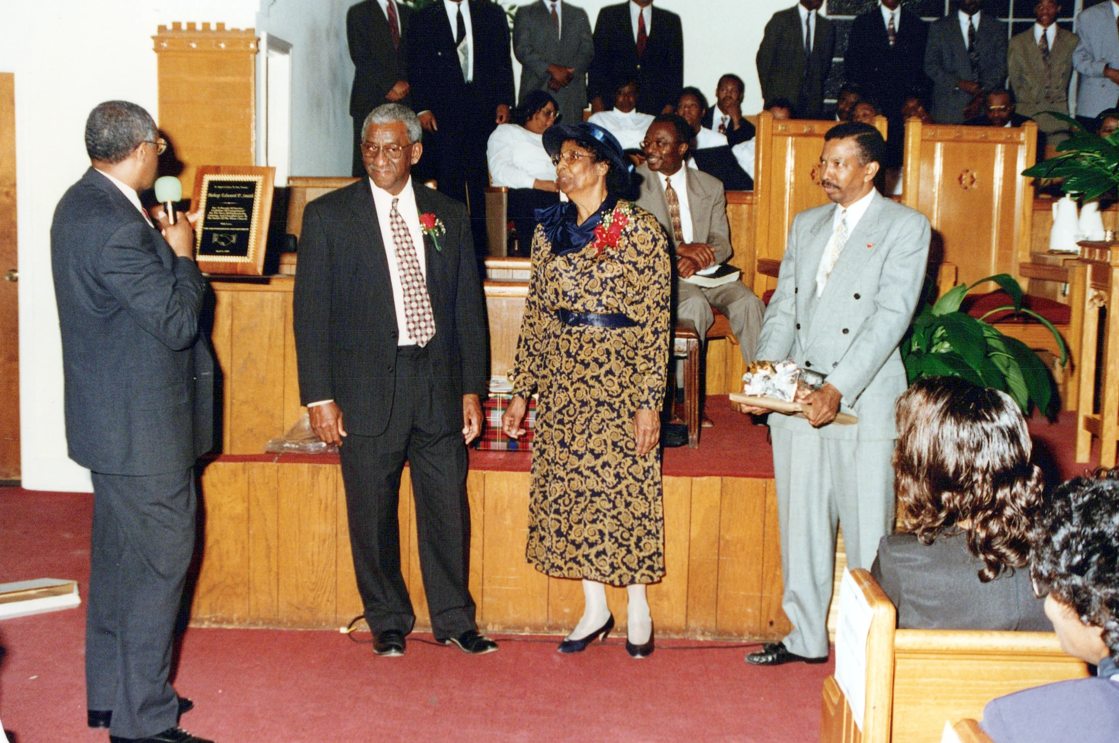 An older Black couple is honored on a church stage. A speaker presents them with a plaque while another man holds a gift and the congregation watches.