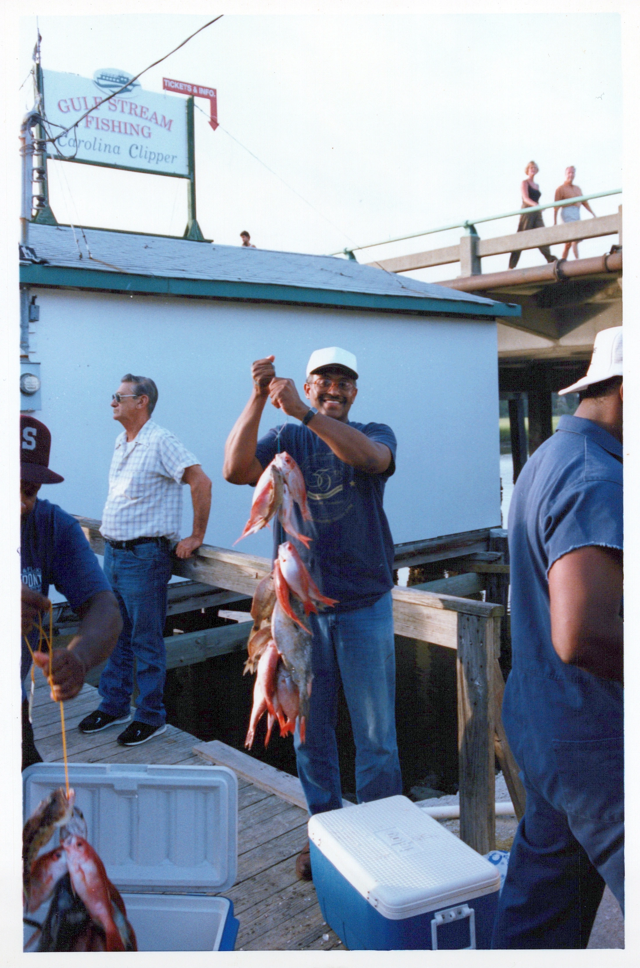 A smiling man in a white cap and glasses proudly holds up a string of red fish he caught, standing on a wooden dock with other men and coolers.
