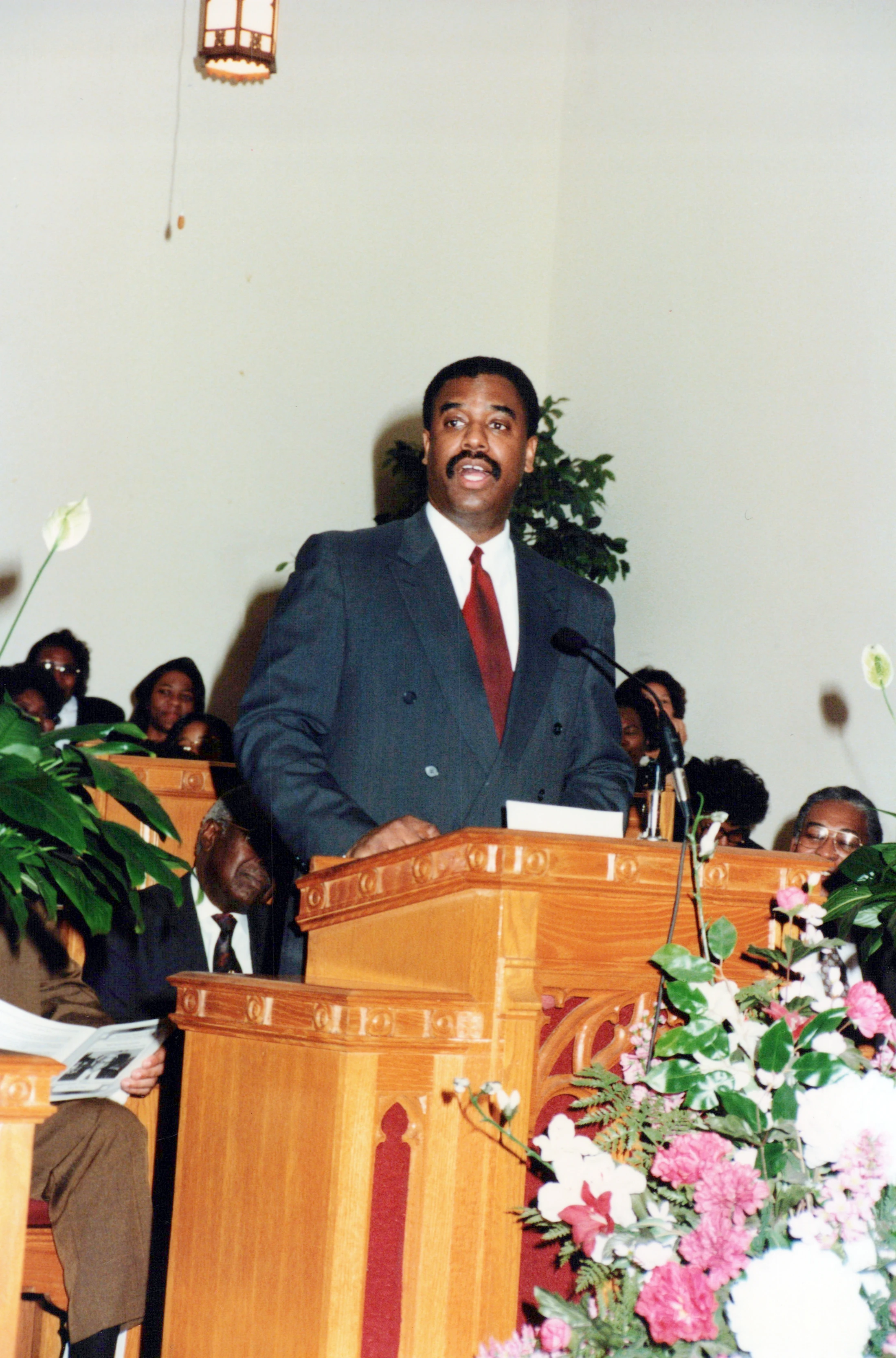A Black man in a dark double-breasted suit and red tie stands at a wooden lectern speaking. A congregation is seated in pews in the background.