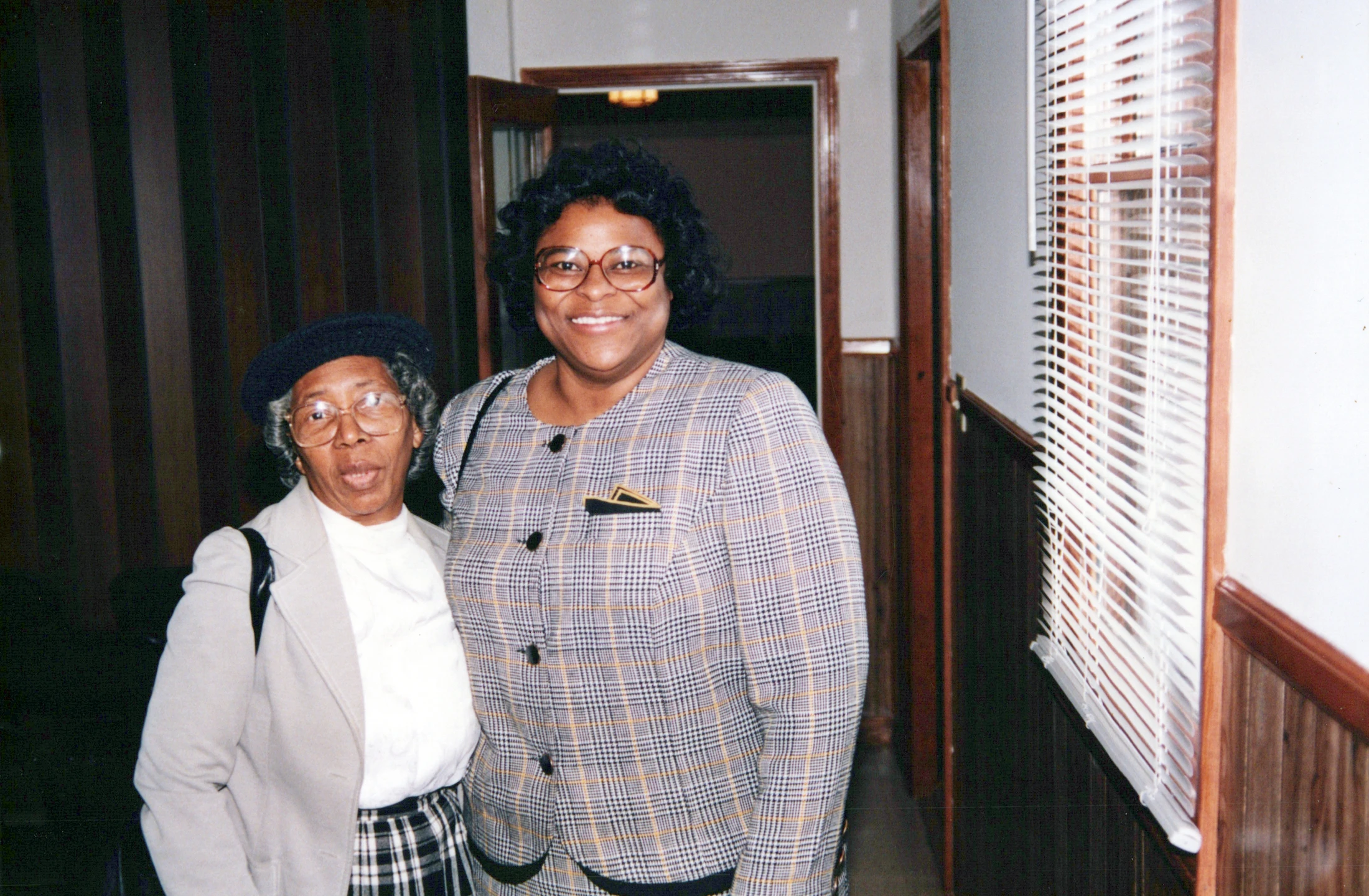 A vintage film photograph of two Black women posing together in a hallway. The woman on the left wears a hat and the woman on the right wears a plaid jacket.