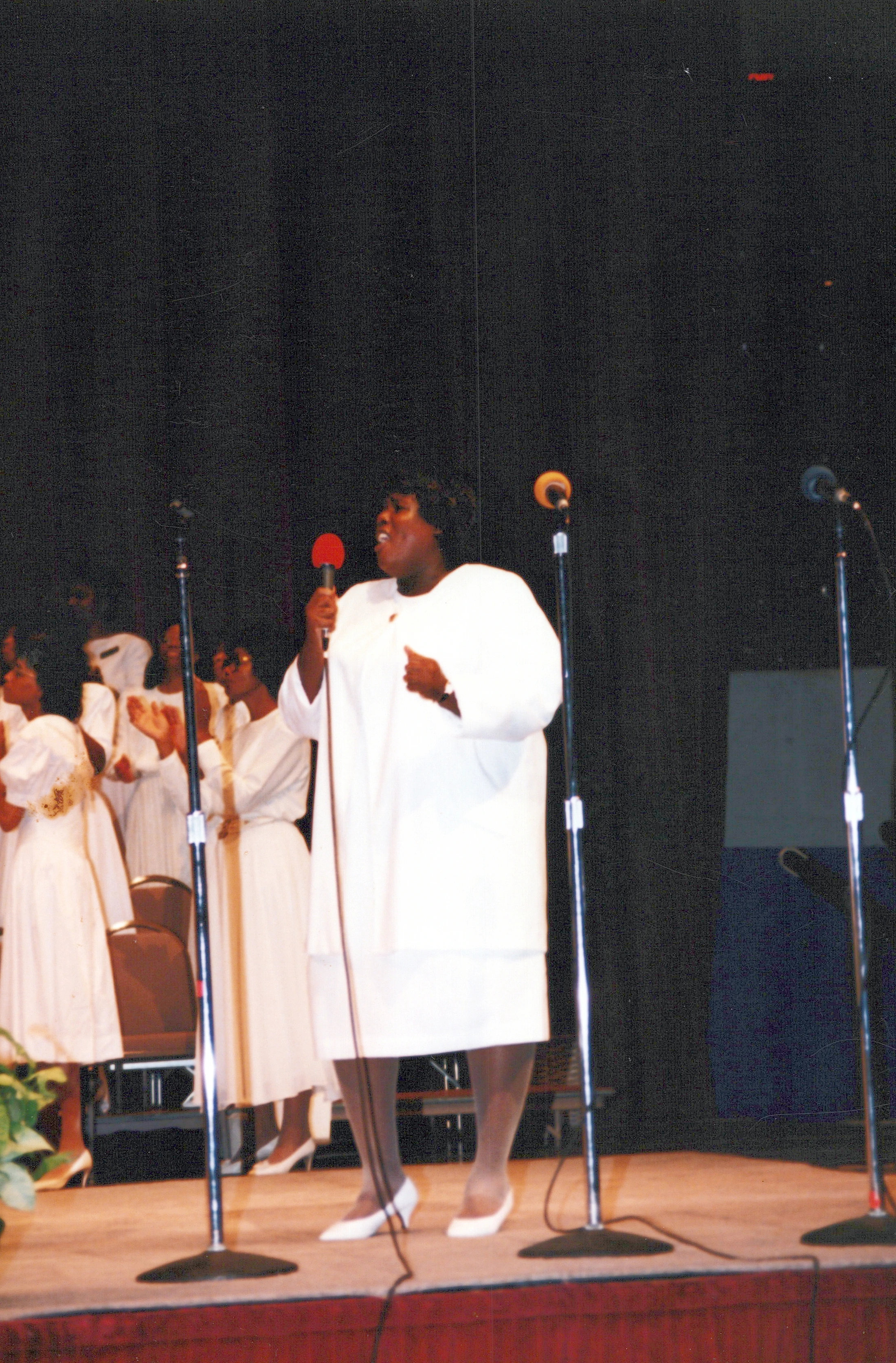 A Black woman in a white dress sings into a microphone on stage. Behind her, a choir, also in white, stands and watches the performance.