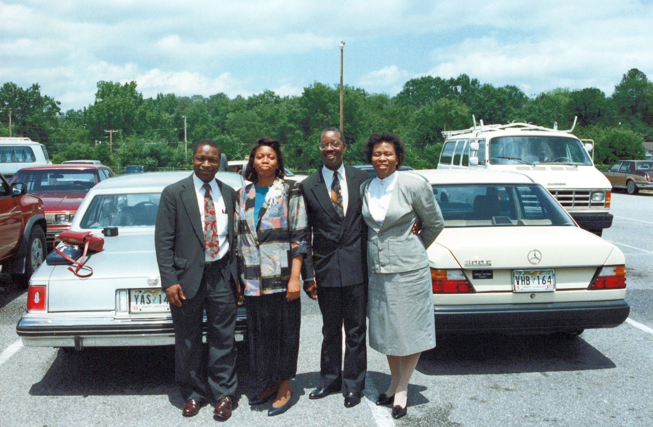 A vintage film photo of two men and two women in formal attire smiling for the camera, standing between two cars in a parking lot on a bright, sunny day.