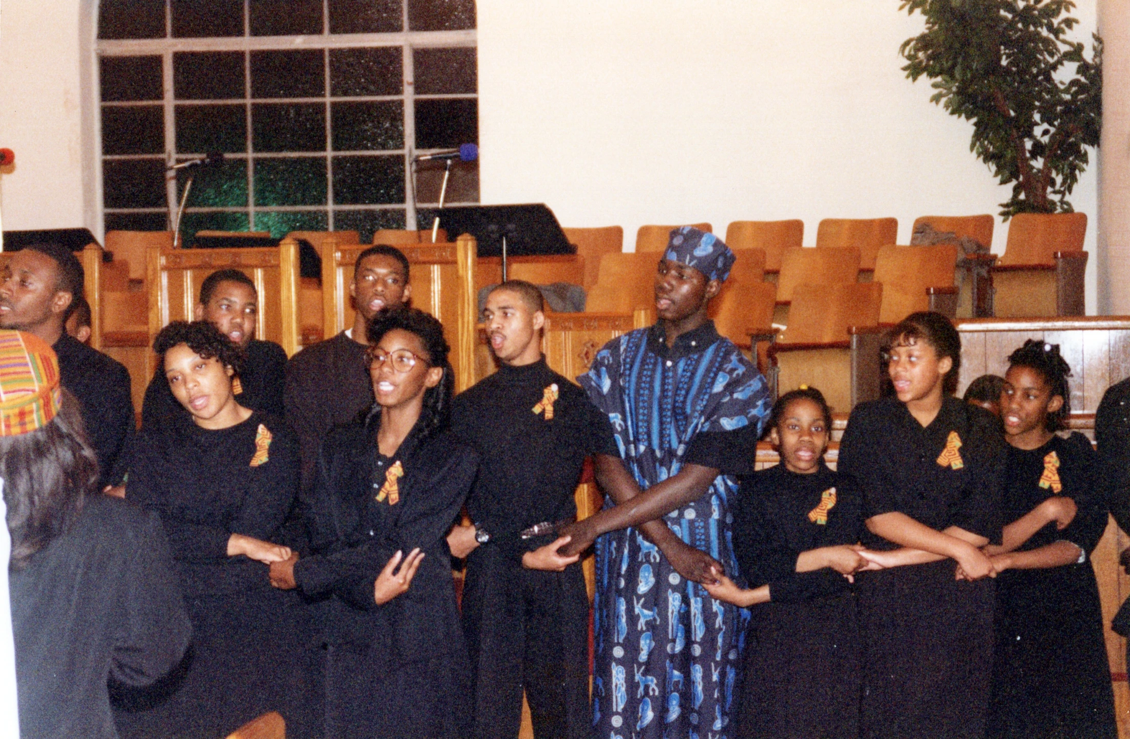 A grainy vintage photograph of a Black choir singing. Most are dressed in black, holding hands. One man wears a blue patterned dashiki and a head wrap.