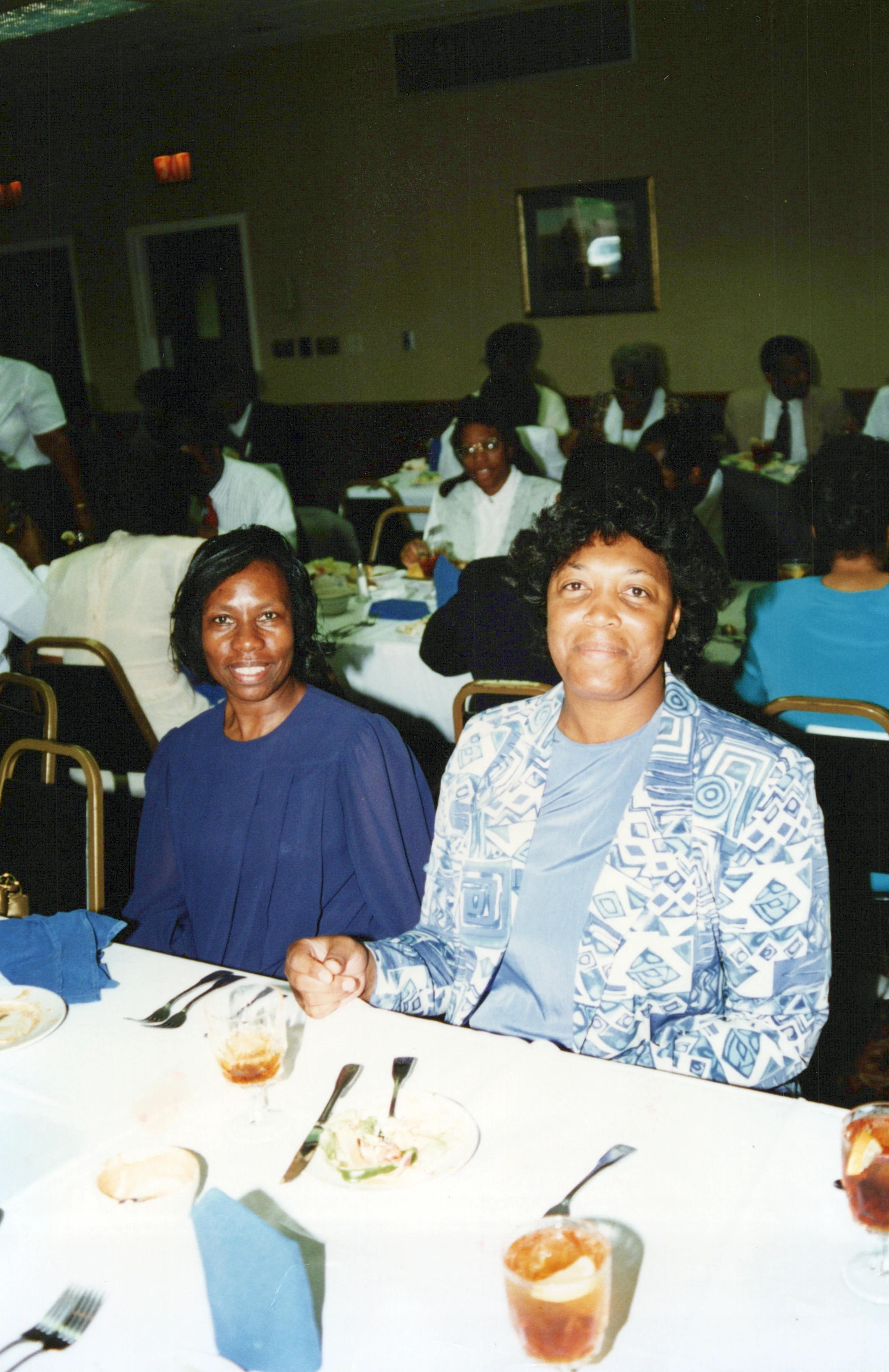 A vintage photograph of two African American women sitting at a dinner table during a banquet. They are both smiling for the camera in a crowded hall.