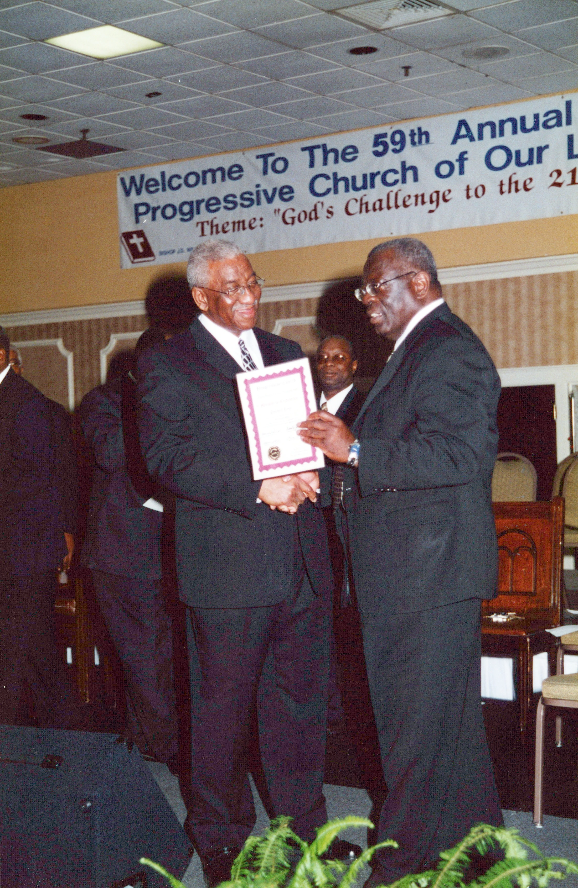 Two older Black men in suits smile as one presents the other with a framed certificate on a stage in front of a banner for a church conference.