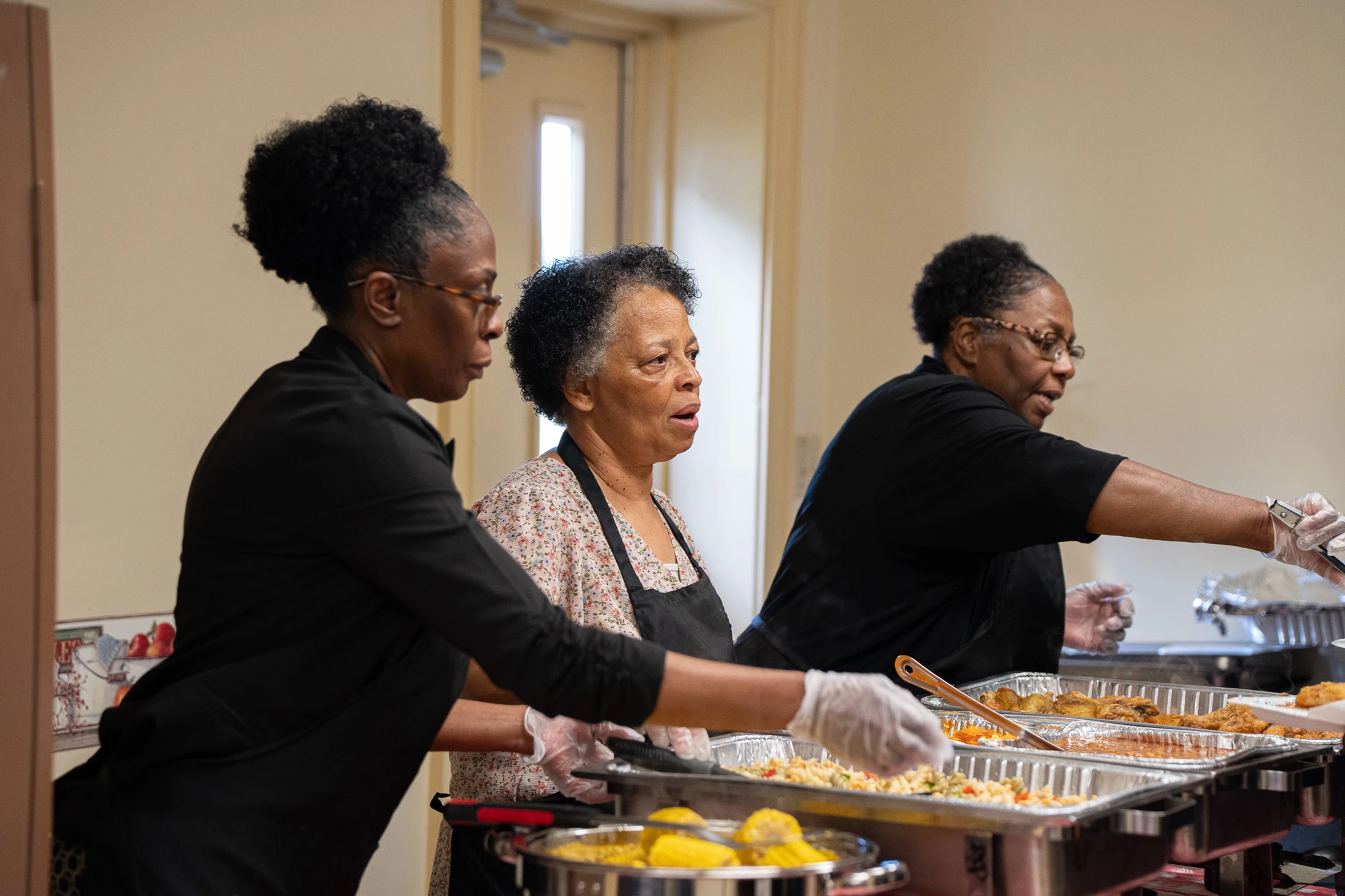 Three Black women stand behind a buffet line with chafing dishes, serving food like corn on the cob and chicken during an indoor gathering.