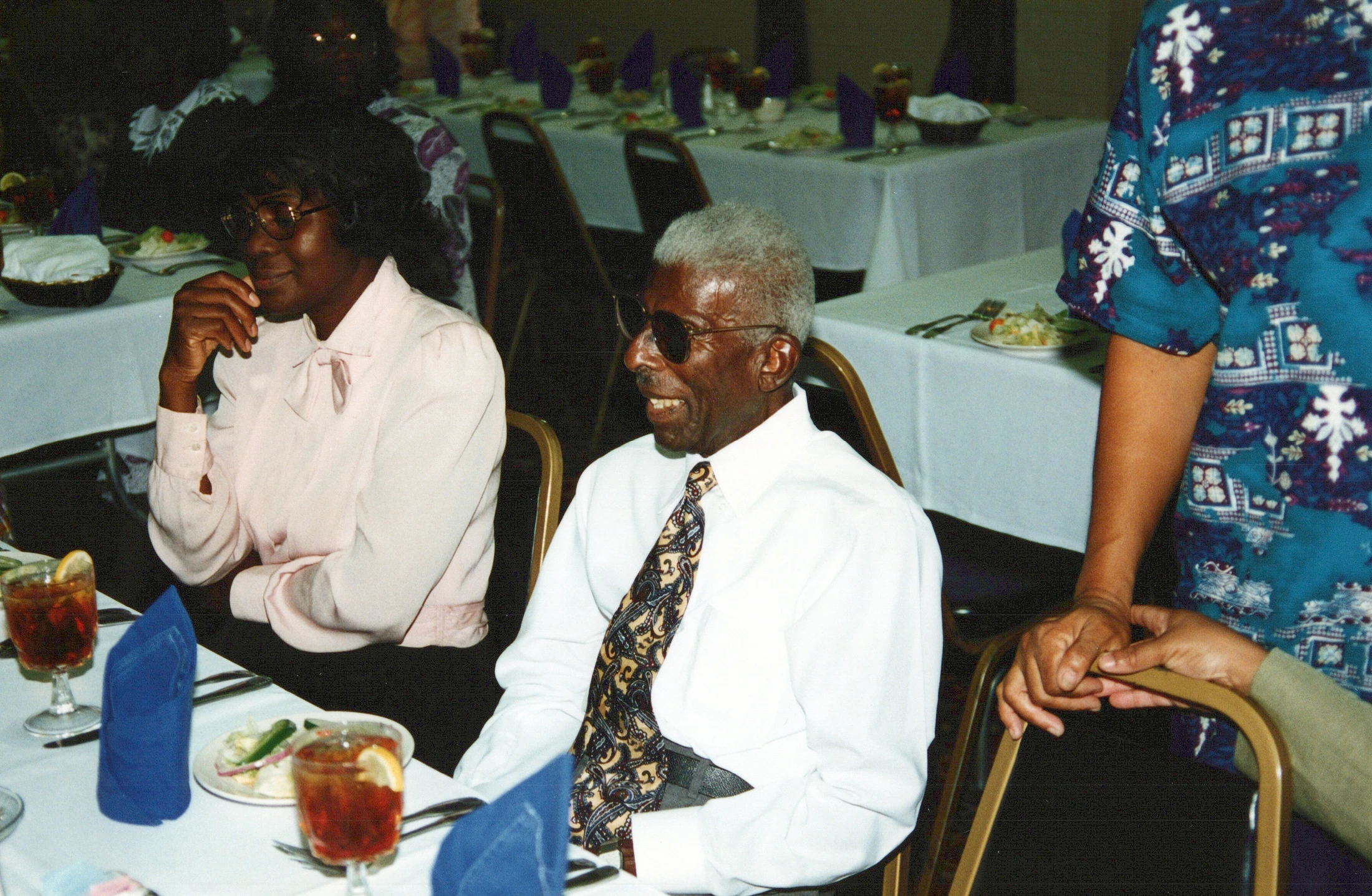 An elderly Black man with grey hair and sunglasses smiles while seated next to a woman in a pink blouse at a formal dinner with white tablecloths.