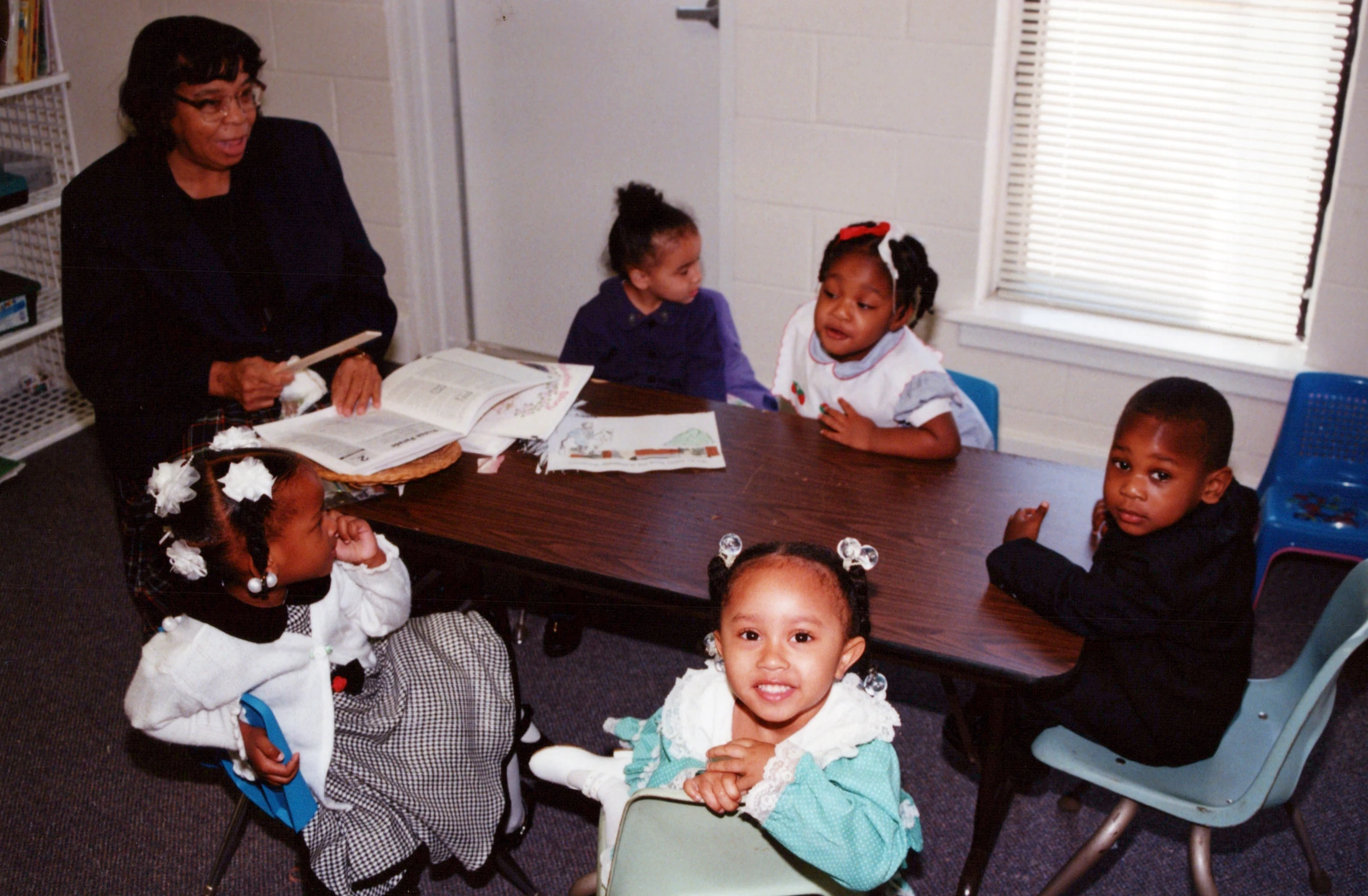 A Black female teacher reads from a book to five young Black children sitting around a table in a classroom. One little girl in front smiles at the camera.