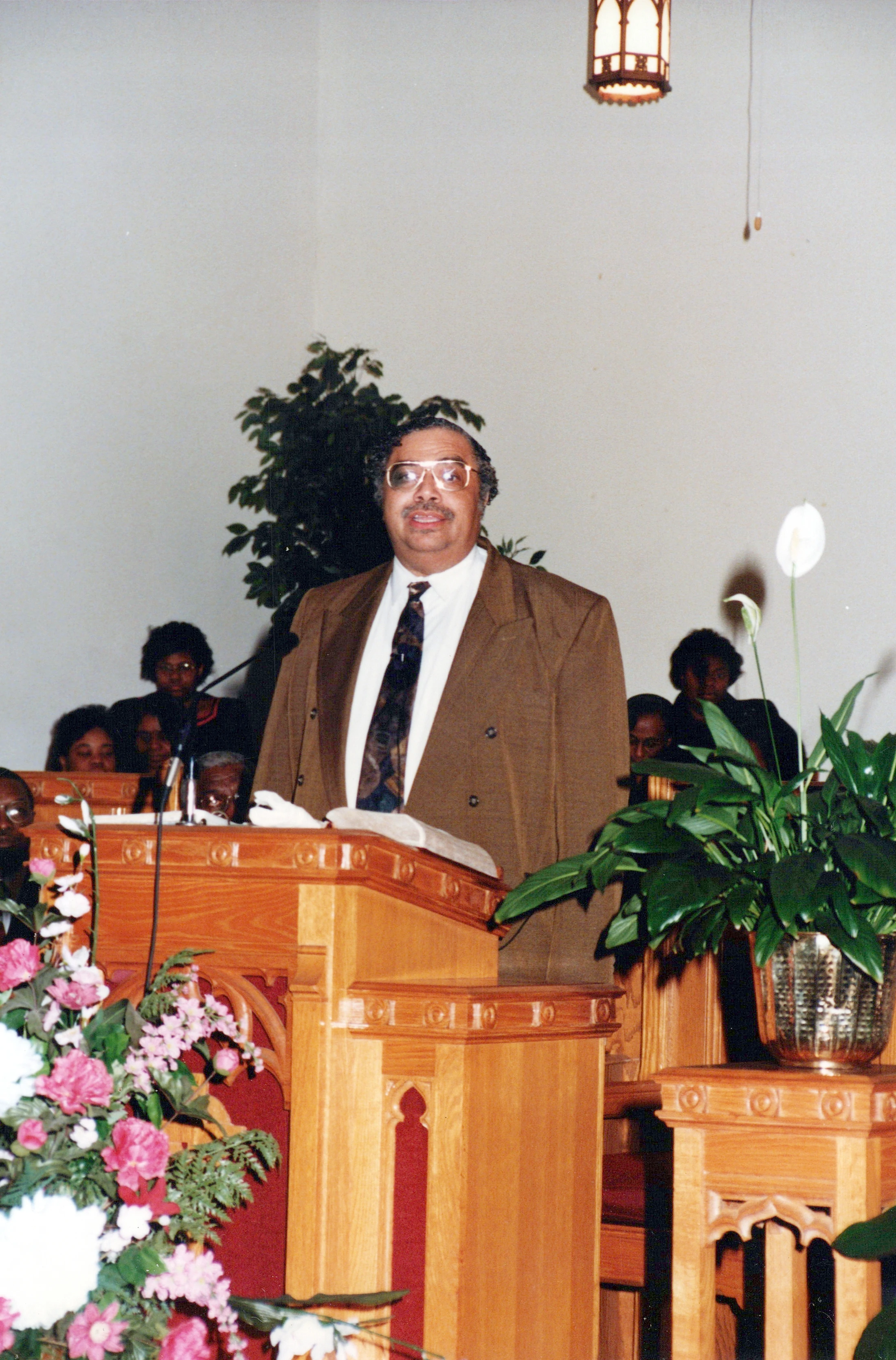 An African American man in a brown suit and glasses smiles as he stands behind a wooden pulpit in a church, with flowers and plants nearby.