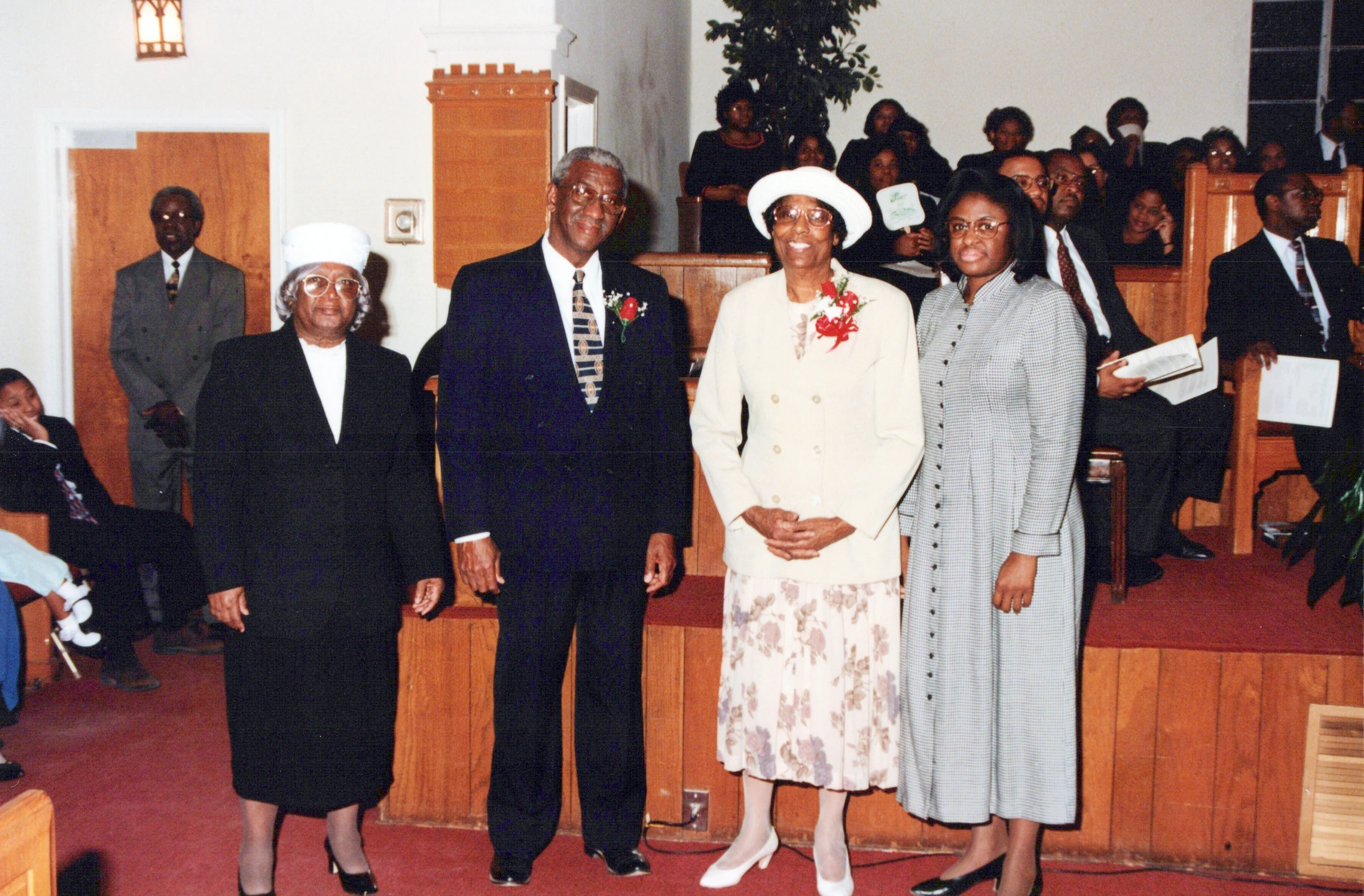 Four older Black adults, two men and two women, stand smiling for a photo at the front of a church, with the congregation seated in pews behind them.