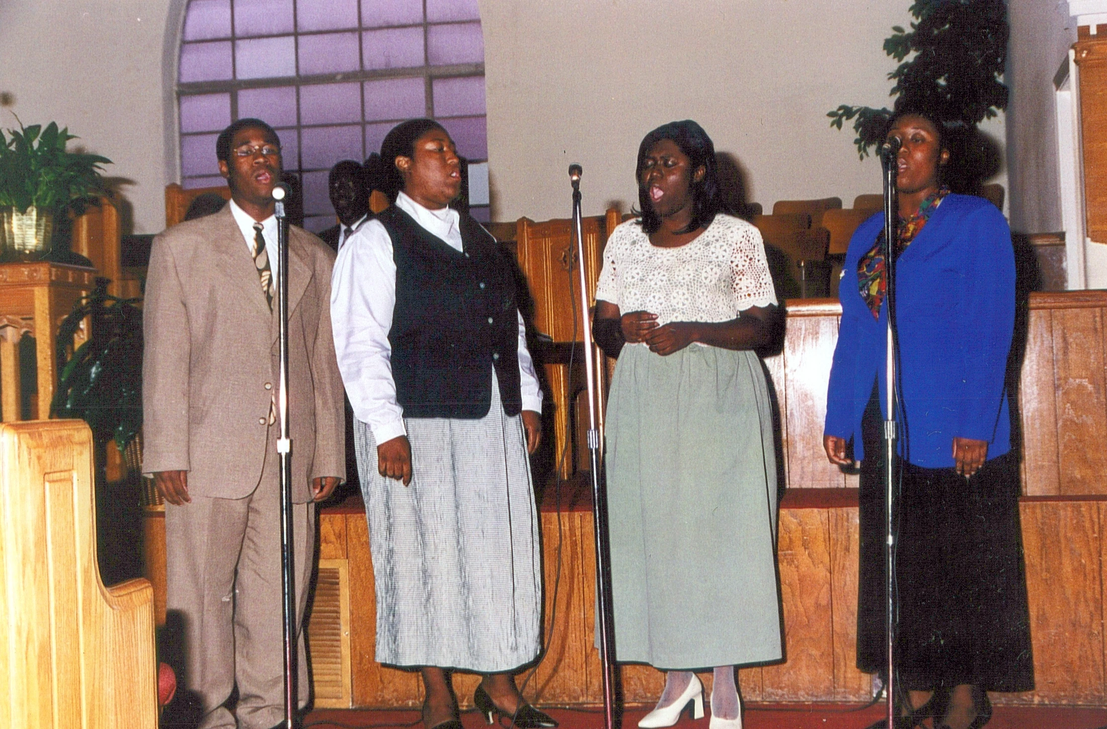 A vintage photograph of four Black singers, one man and three women, standing in a line at microphones and singing in a church sanctuary with wooden pews.