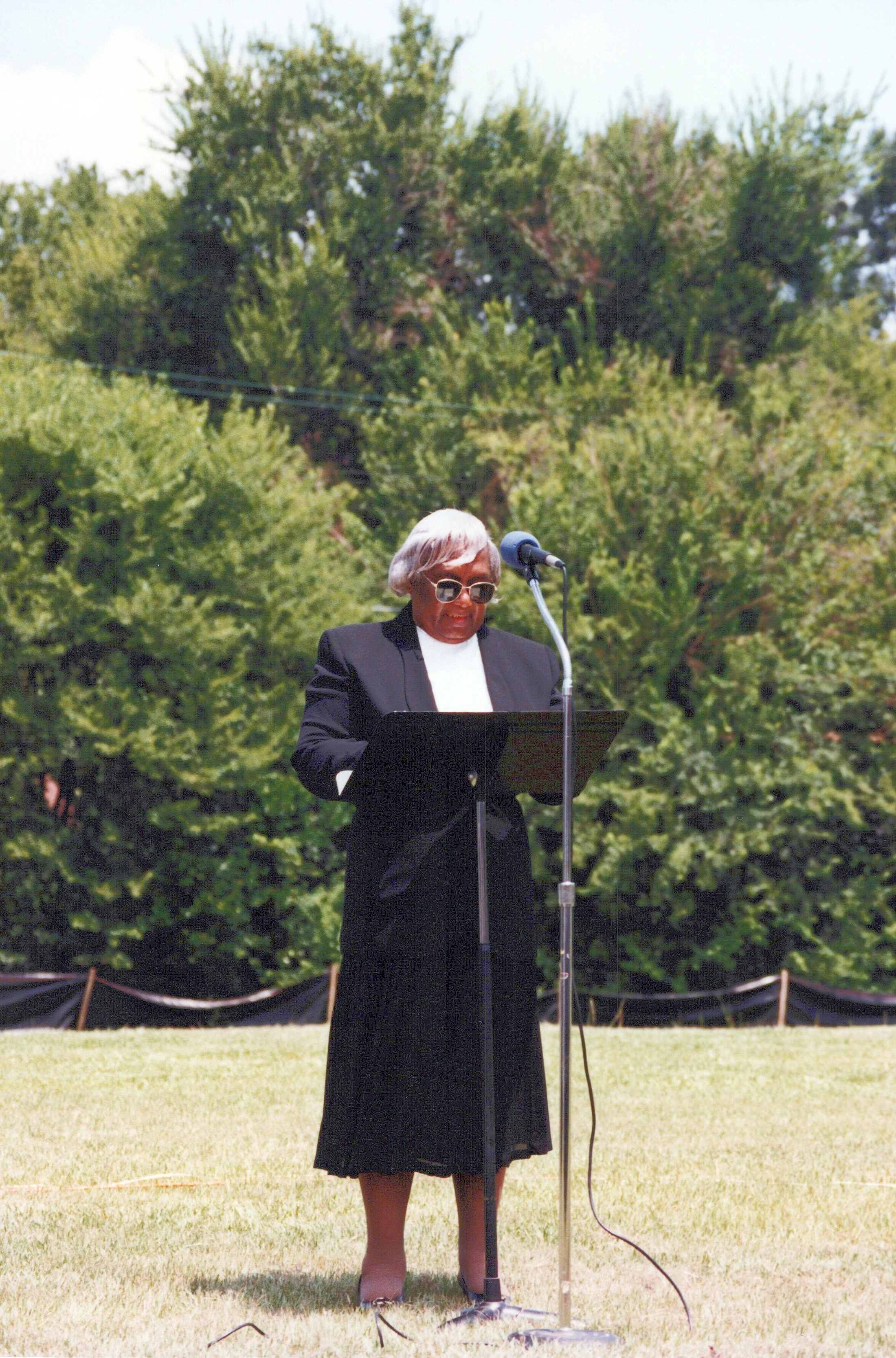 An elderly African American woman with white hair and sunglasses speaks at a podium outdoors. She stands on a grassy lawn with trees in the background.