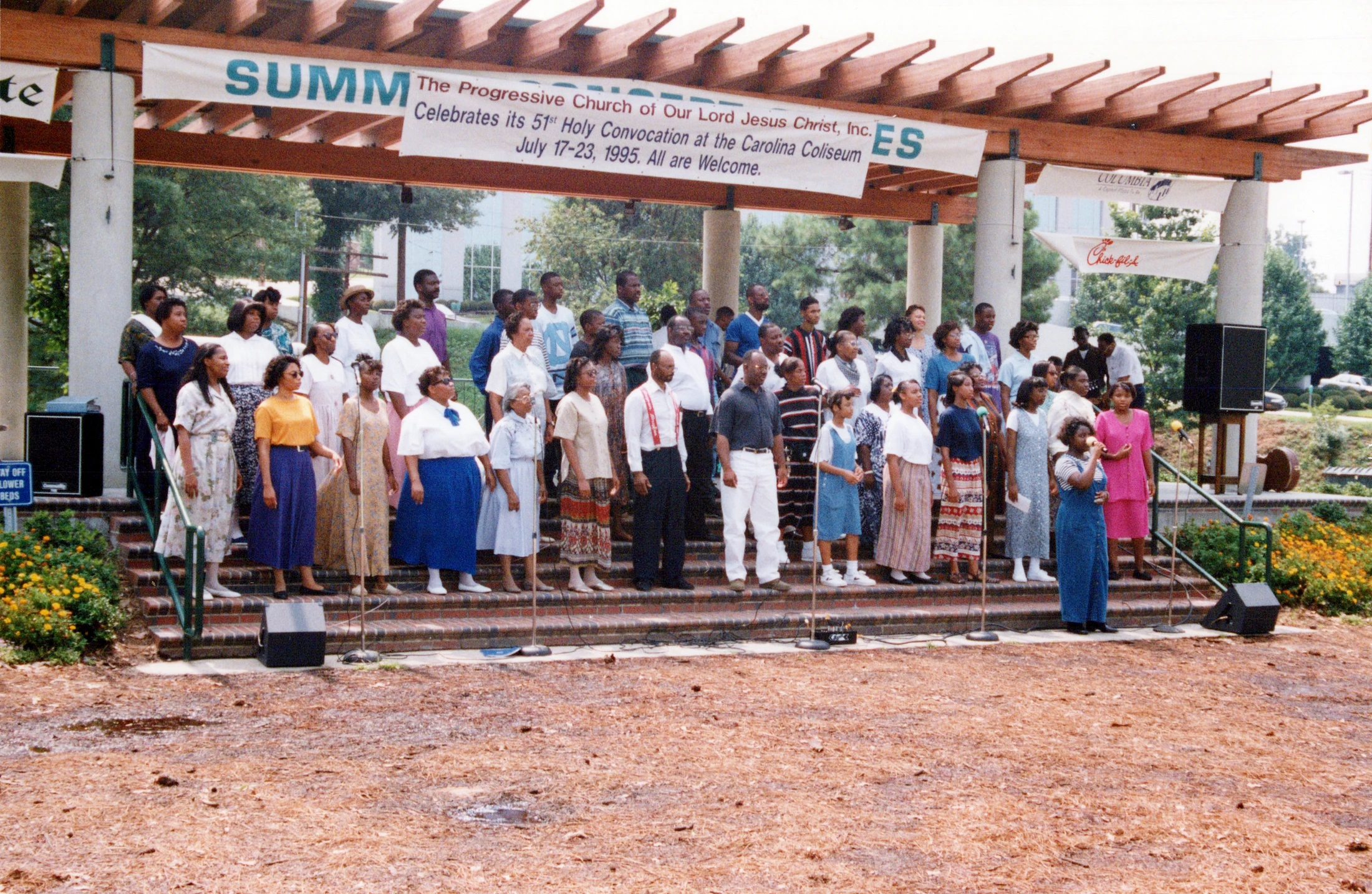 A large church choir of Black men and women stands on steps under a wooden pergola, performing outdoors for a Holy Convocation event in July 1995.