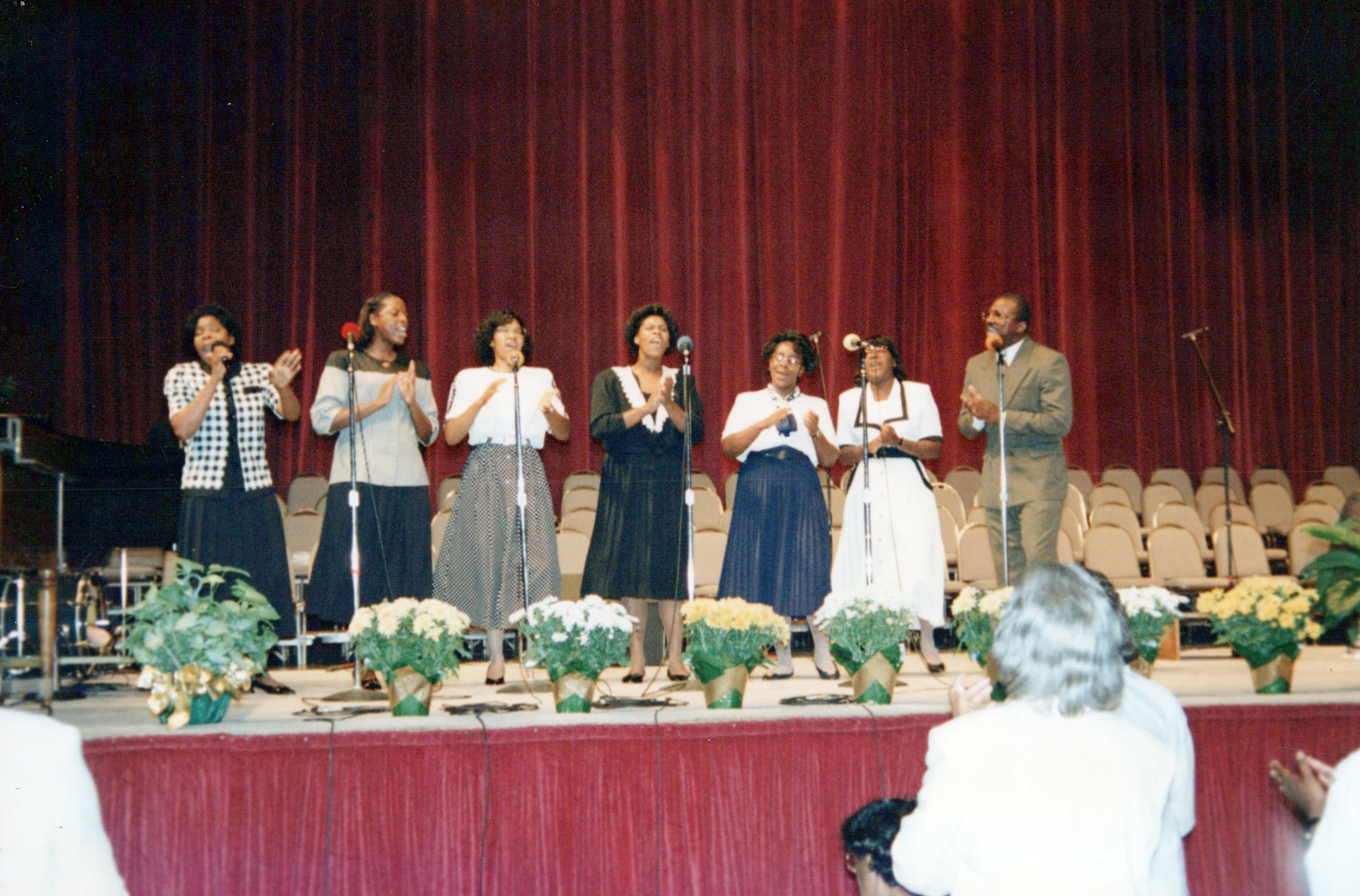 A group of seven African American singers, six women and one man, perform on a stage with a red curtain. They stand at microphones, singing and clapping.