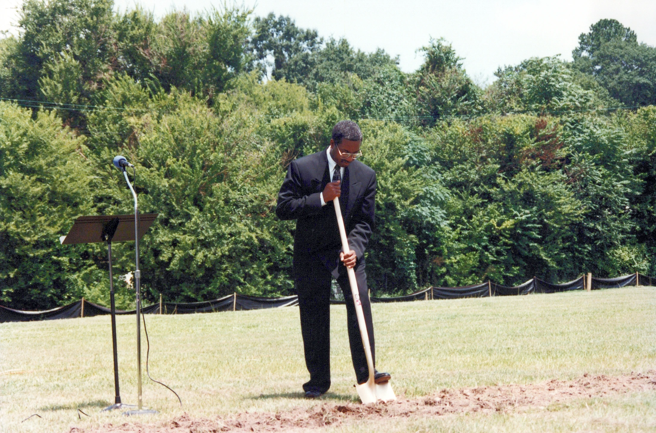 An African American man in a black suit and tie digs with a shovel in a grassy field during a groundbreaking ceremony. A microphone on a stand is to his left.