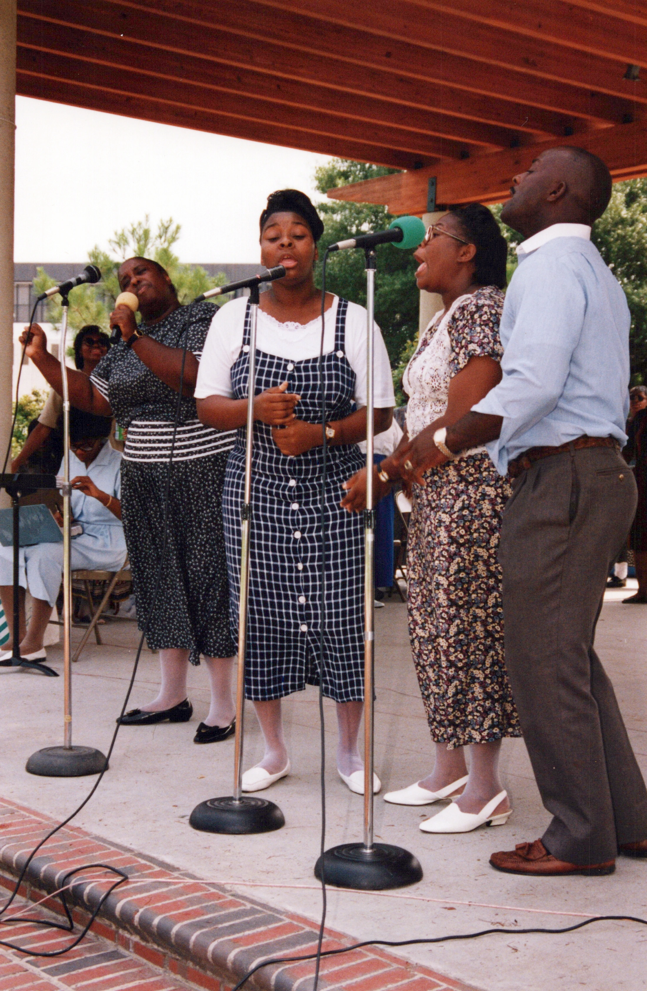 A group of four Black singers, three women and one man, stand in a line on an outdoor stage, singing passionately into microphones.