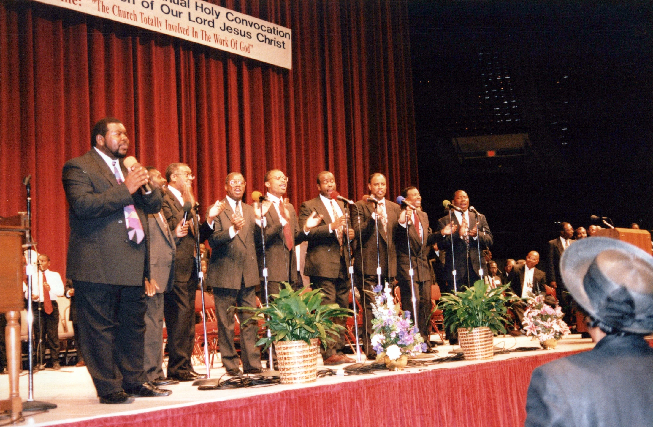 A group of Black men in dark suits sing into microphones on a stage with a red curtain backdrop during a church convocation.
