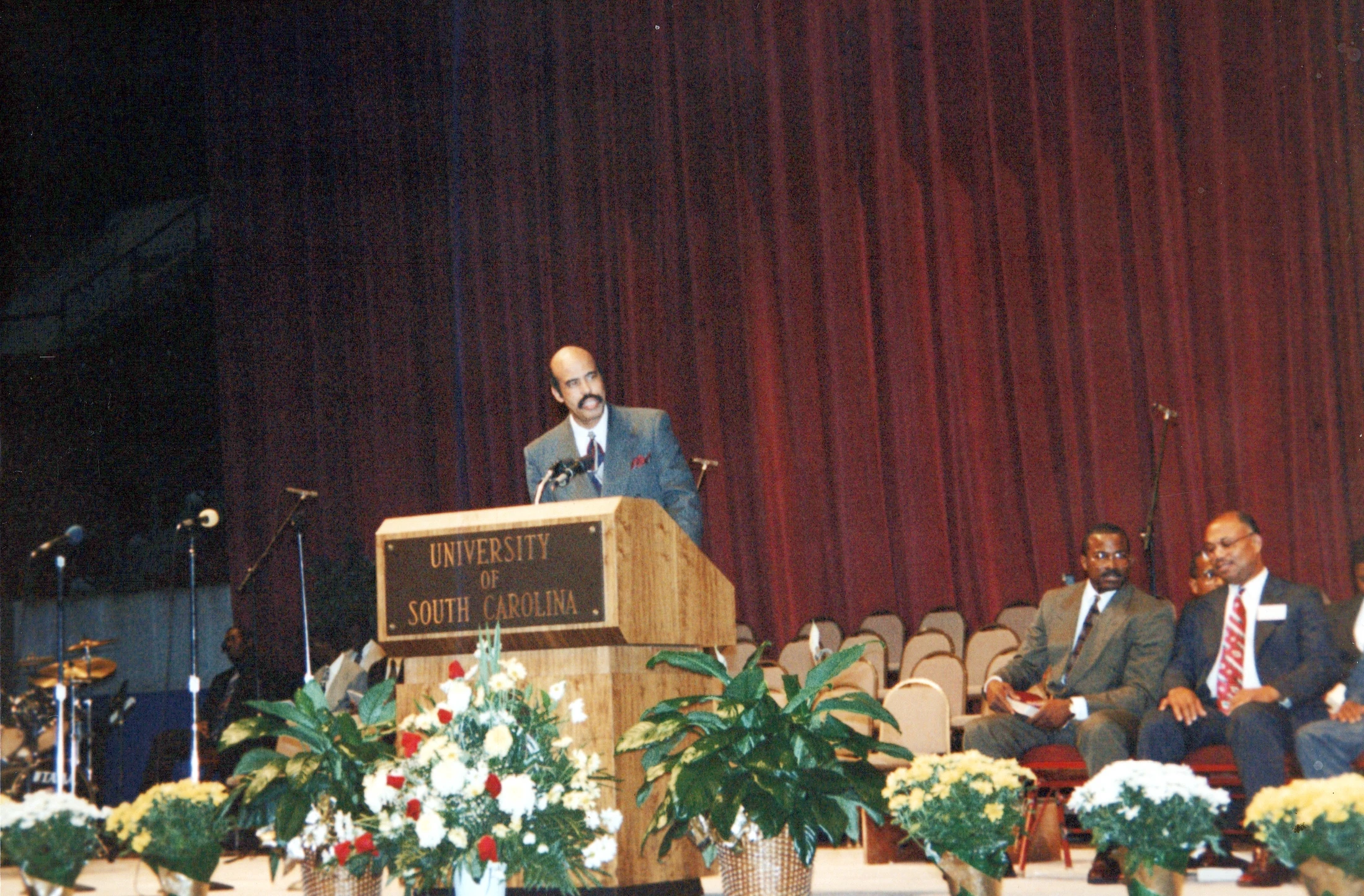 A Black man in a suit with a mustache speaks at a wooden podium bearing the University of South Carolina name. Other men are seated on stage behind him.