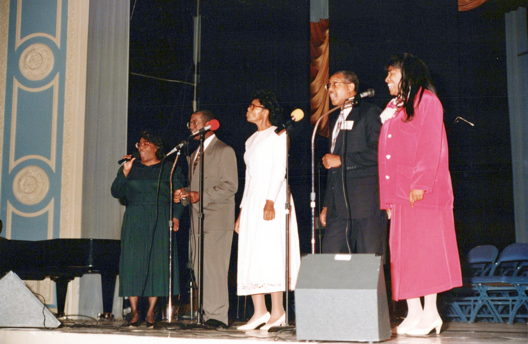 A vintage photograph shows five African American singers, three women and two men, standing in a line on a stage and singing into microphones.