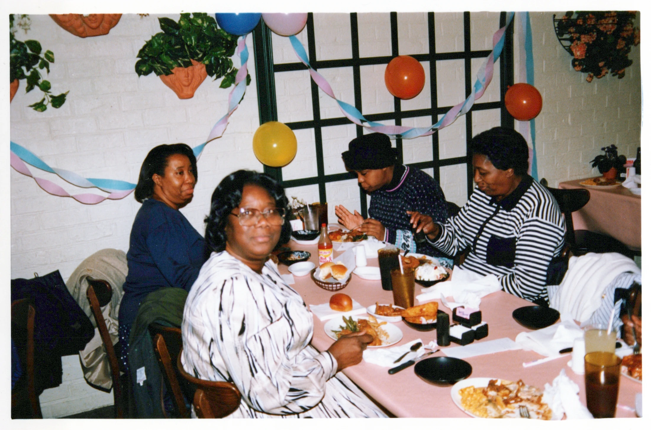 A vintage photograph shows four Black women seated at a dinner table decorated with balloons and streamers, enjoying a meal together at a celebration.