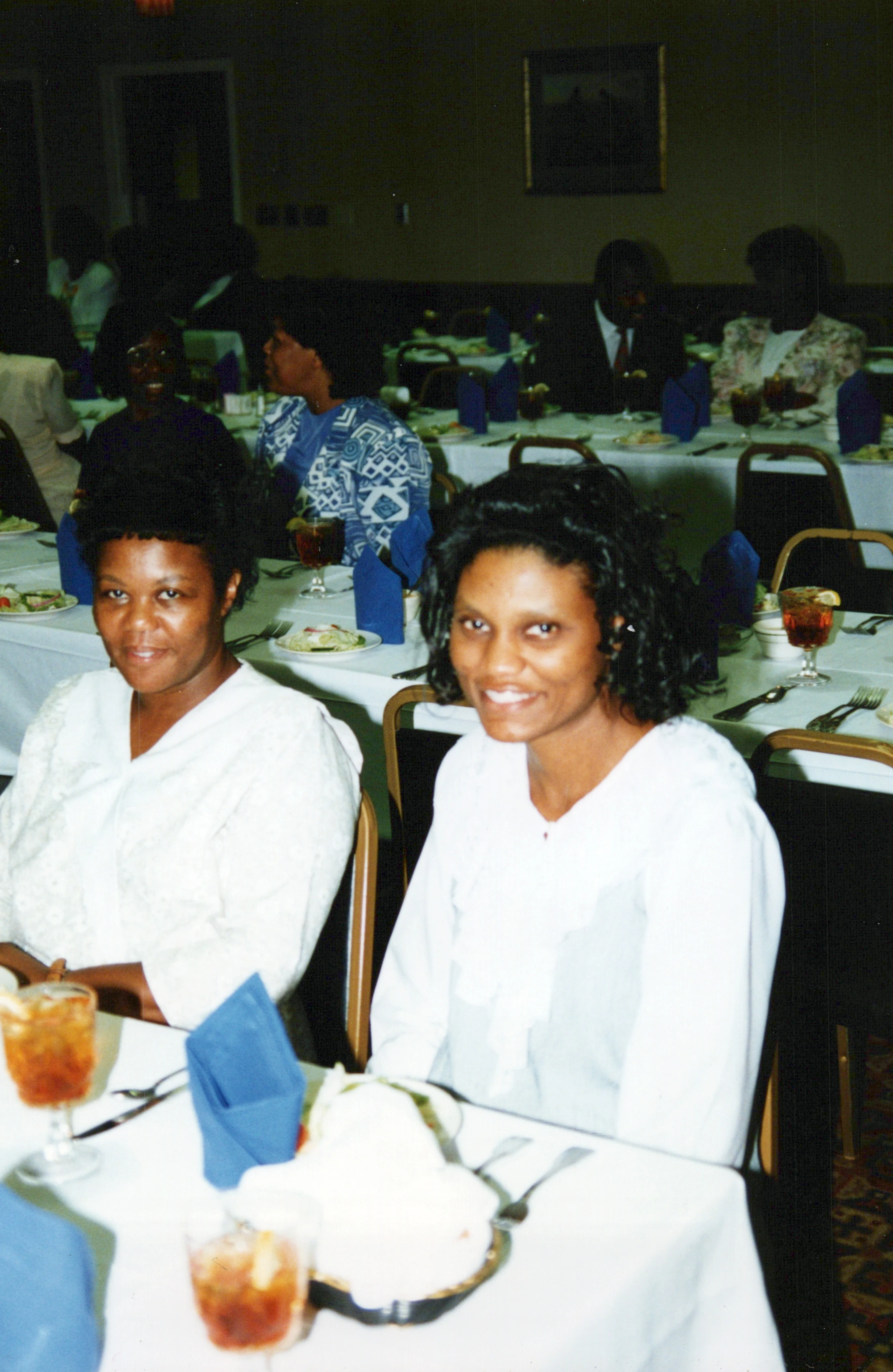A vintage photograph of two Black women smiling at the camera while seated at a table during a formal dinner or banquet, with other guests in the background.