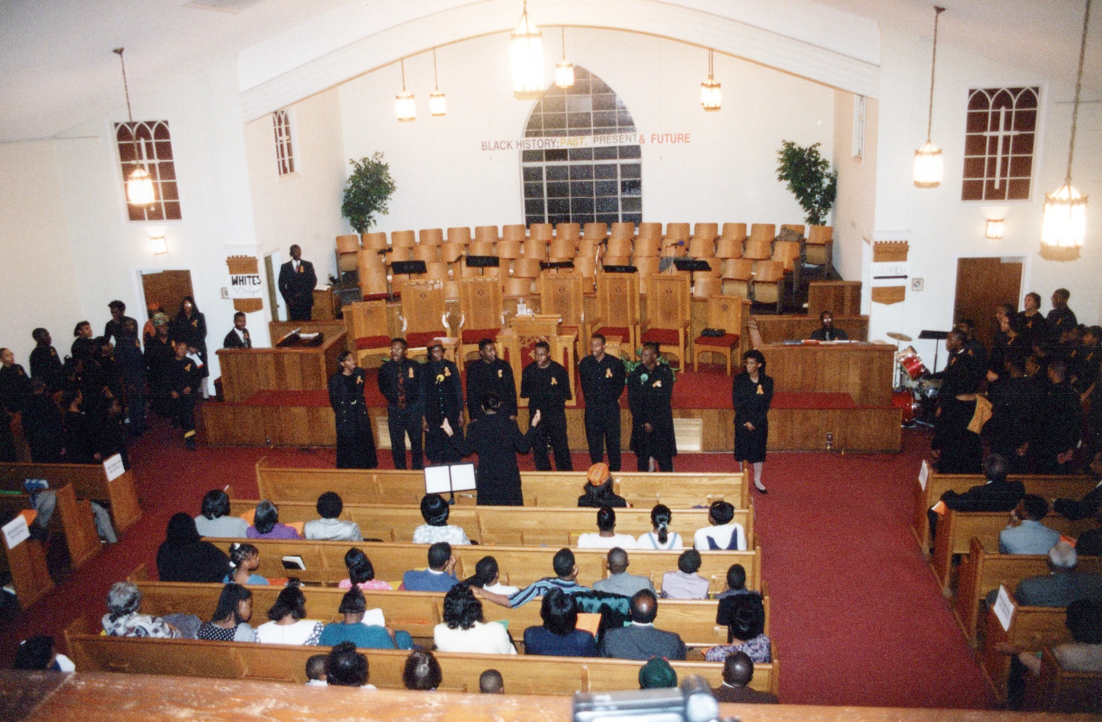 A choir performs for a congregation inside a church during a Black History event, with historical segregation signs displayed as part of the performance.