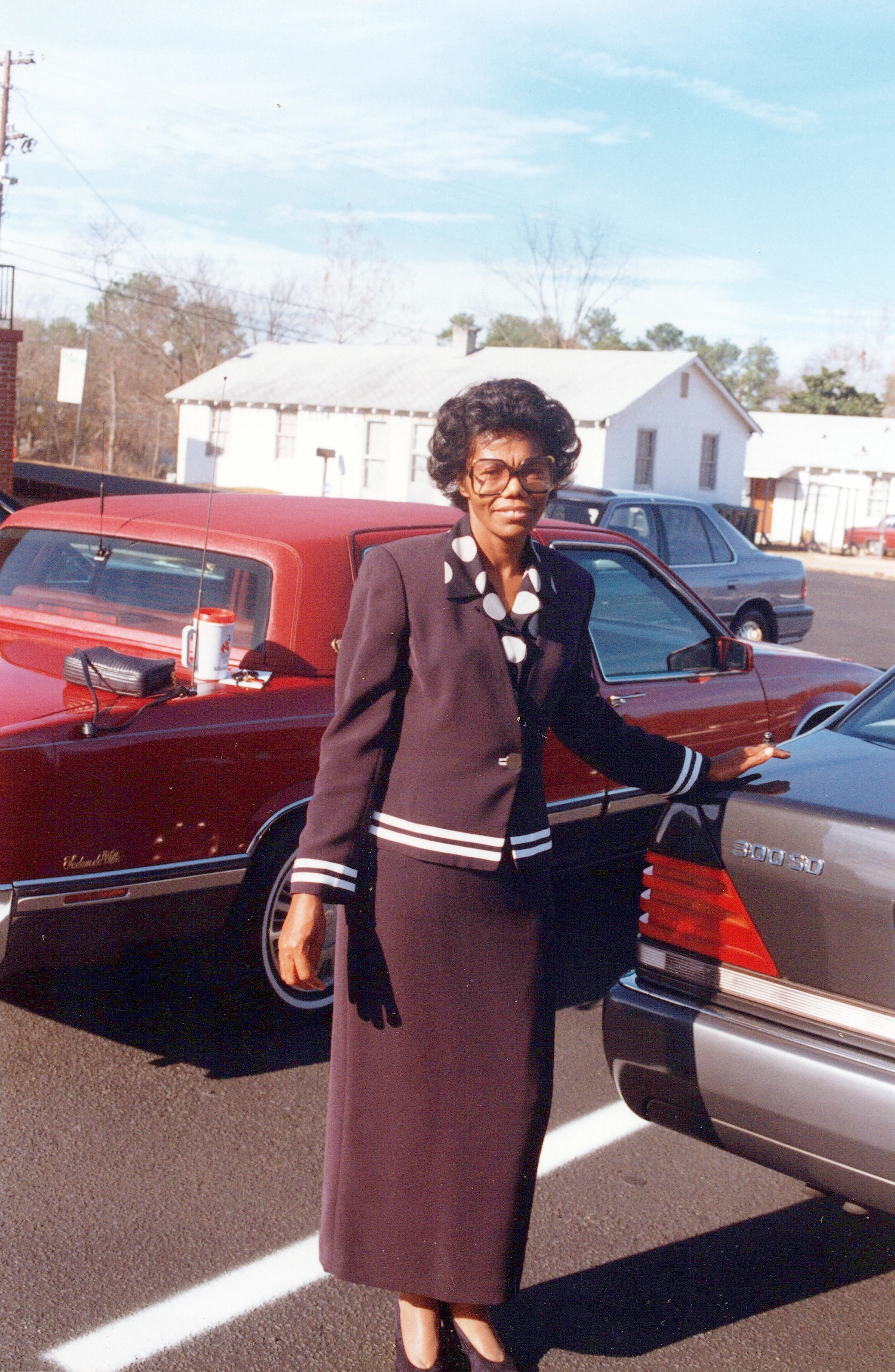 A full-length photo of a Black woman in a dark skirt suit and glasses, smiling as she stands between a red car and a gray car in a parking lot.