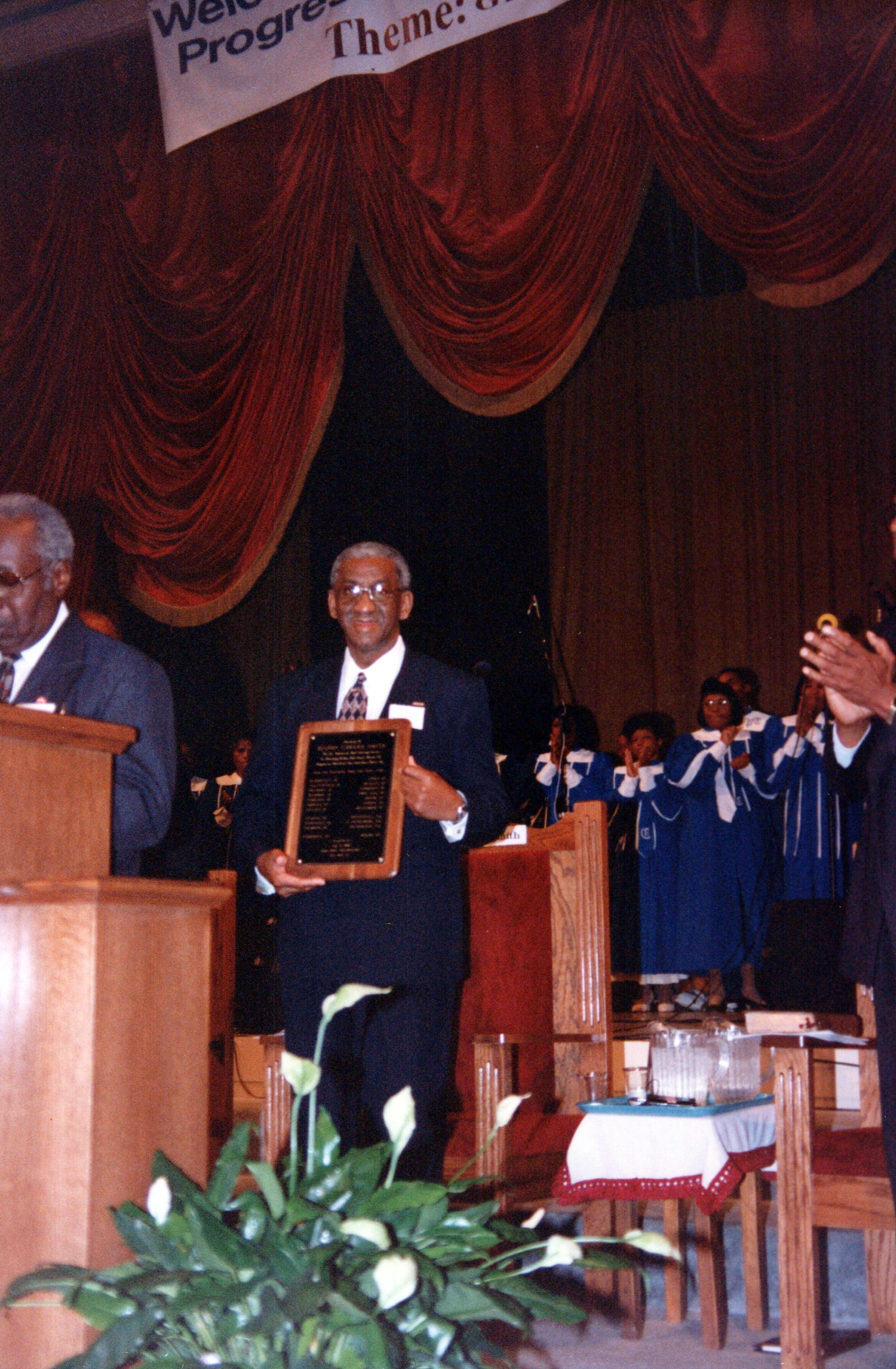 An older Black man in a suit and glasses smiles as he holds a wooden plaque on a stage. Behind him, a choir in blue and white robes claps.