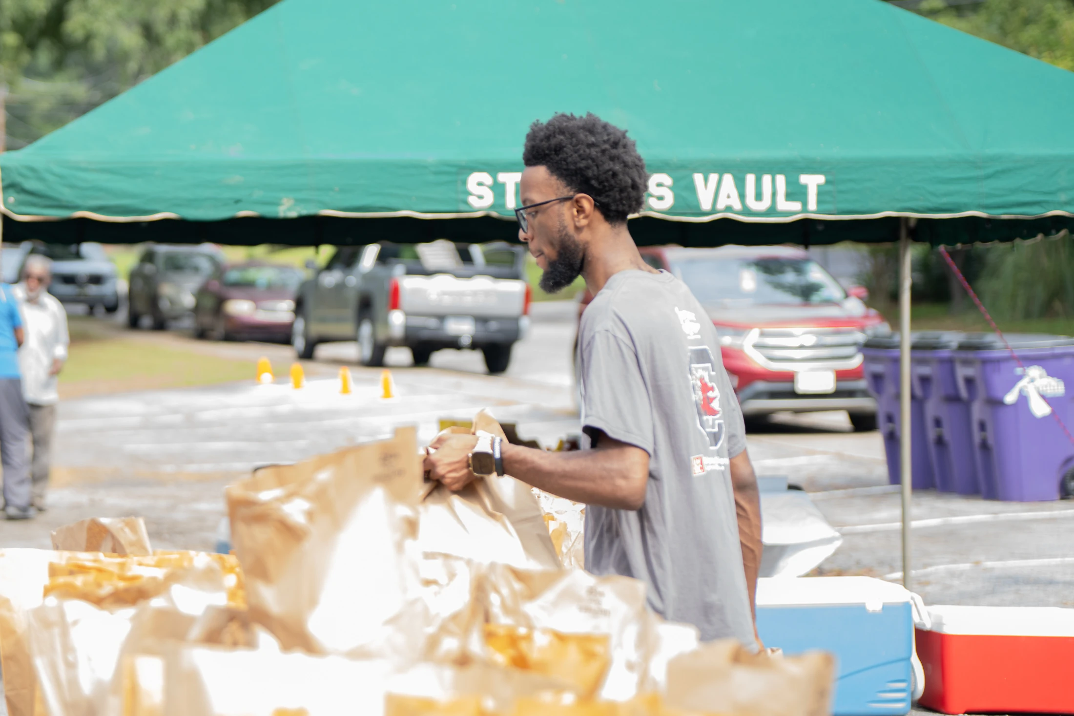 A young Black man with glasses and a beard stands under a green tent, sorting through brown paper bags at a drive-thru community giveaway event.