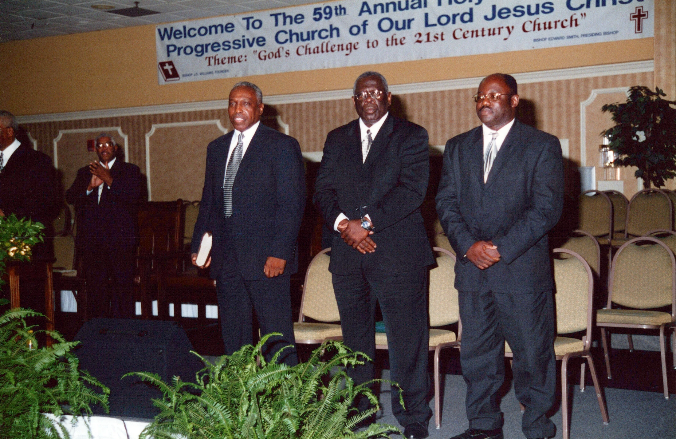 Four African American men in dark suits stand in front of a banner for the 59th Annual Holy Progressive Church of Our Lord Jesus Christ.