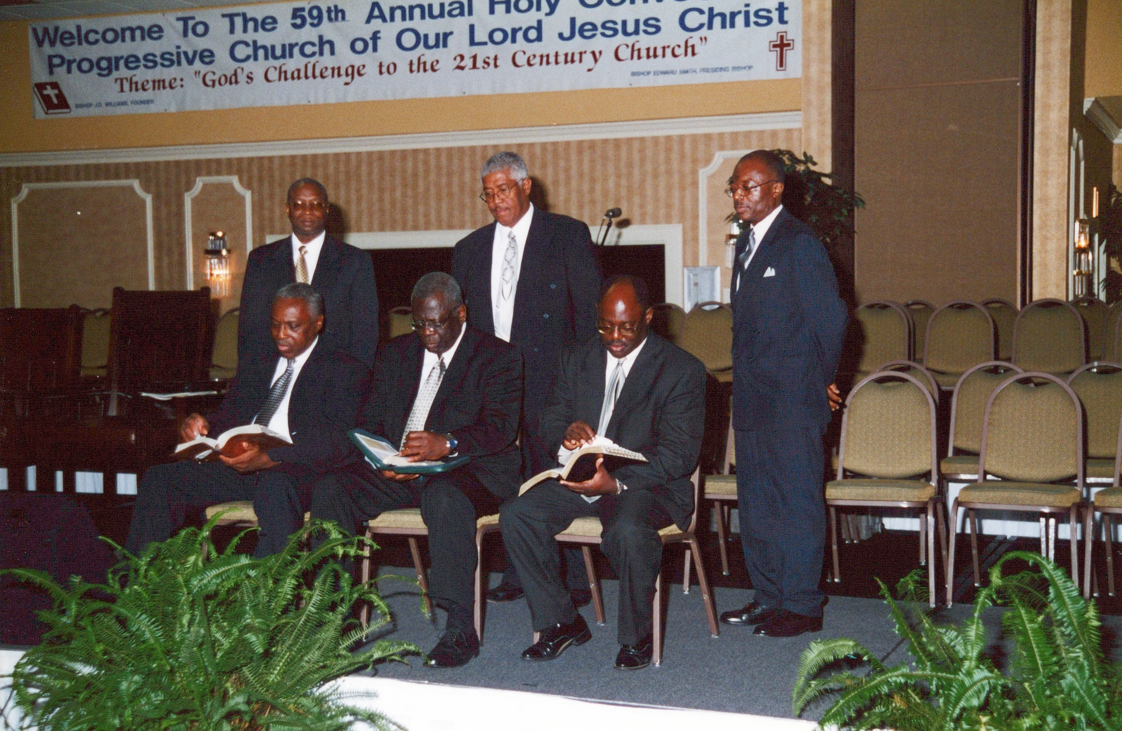 Six African American men in suits on a stage at a church convention. Three are seated reading books, and three stand behind them under a large banner.