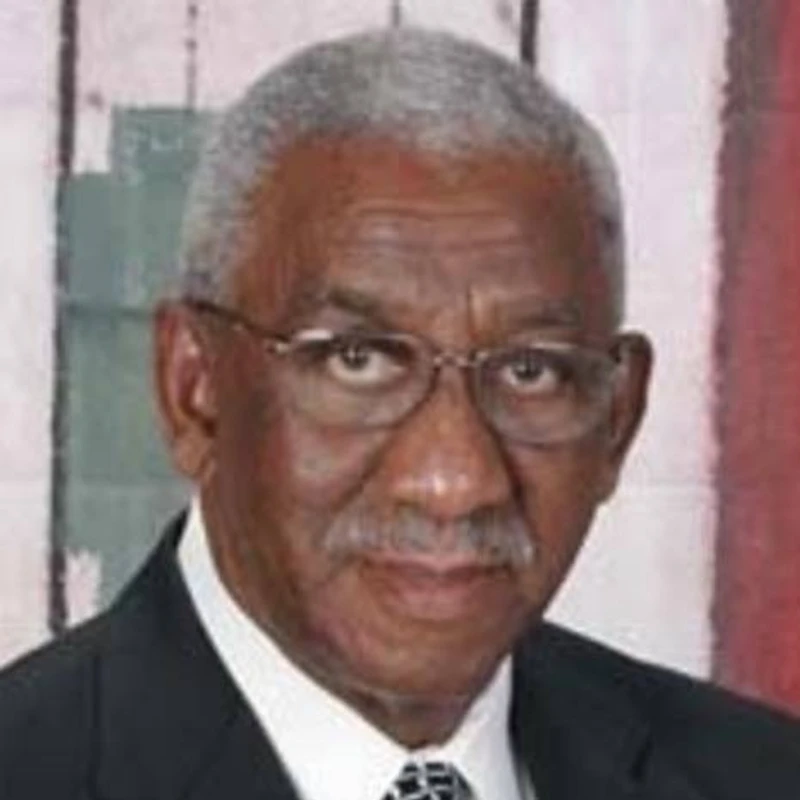 A headshot of an older Black man with gray hair and a mustache, wearing glasses, a dark suit, a white shirt, and a patterned tie.