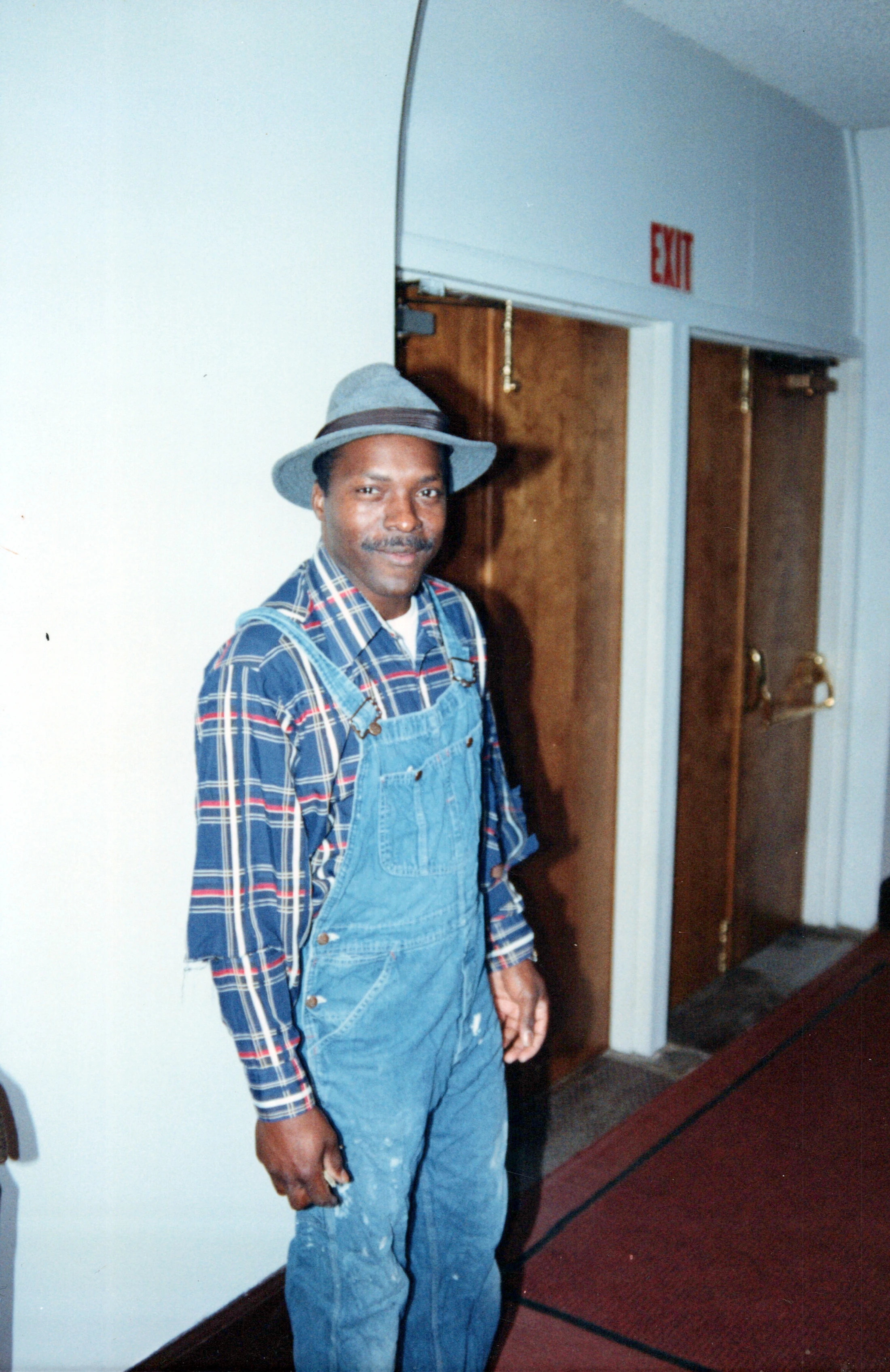 A Black man with a mustache wearing a grey fedora, plaid shirt, and denim overalls smiles while standing in a hallway with an EXIT sign in the background.