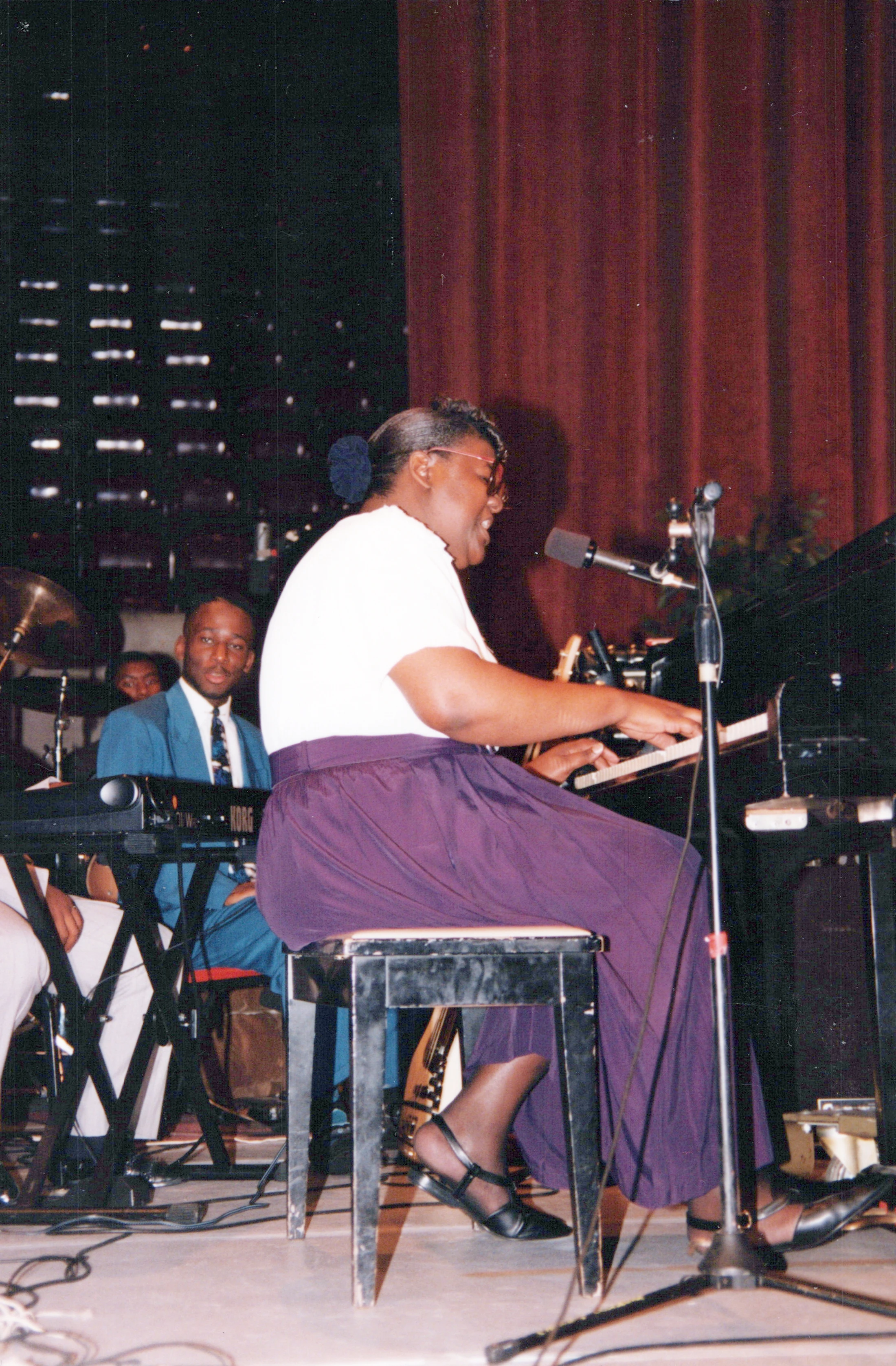 A woman in a white shirt and purple skirt sits at a piano on stage, playing and singing into a microphone. A man in a blue suit is in the background.