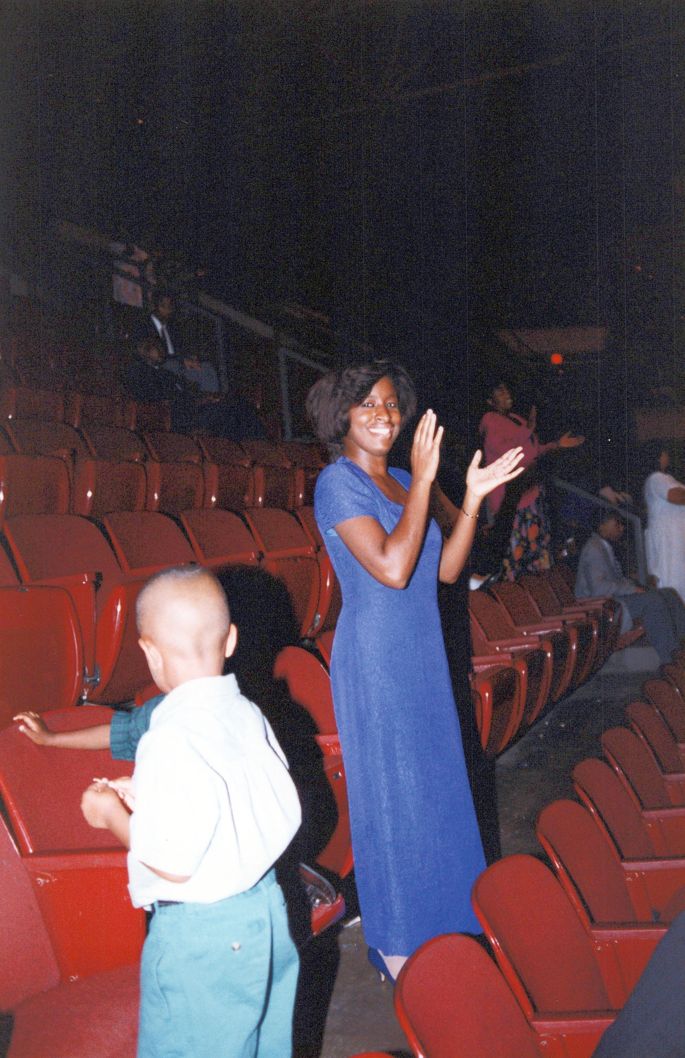 A grainy, vintage photograph of a Black woman in a blue dress smiling and clapping in an auditorium with red seats, with a small boy in the foreground.