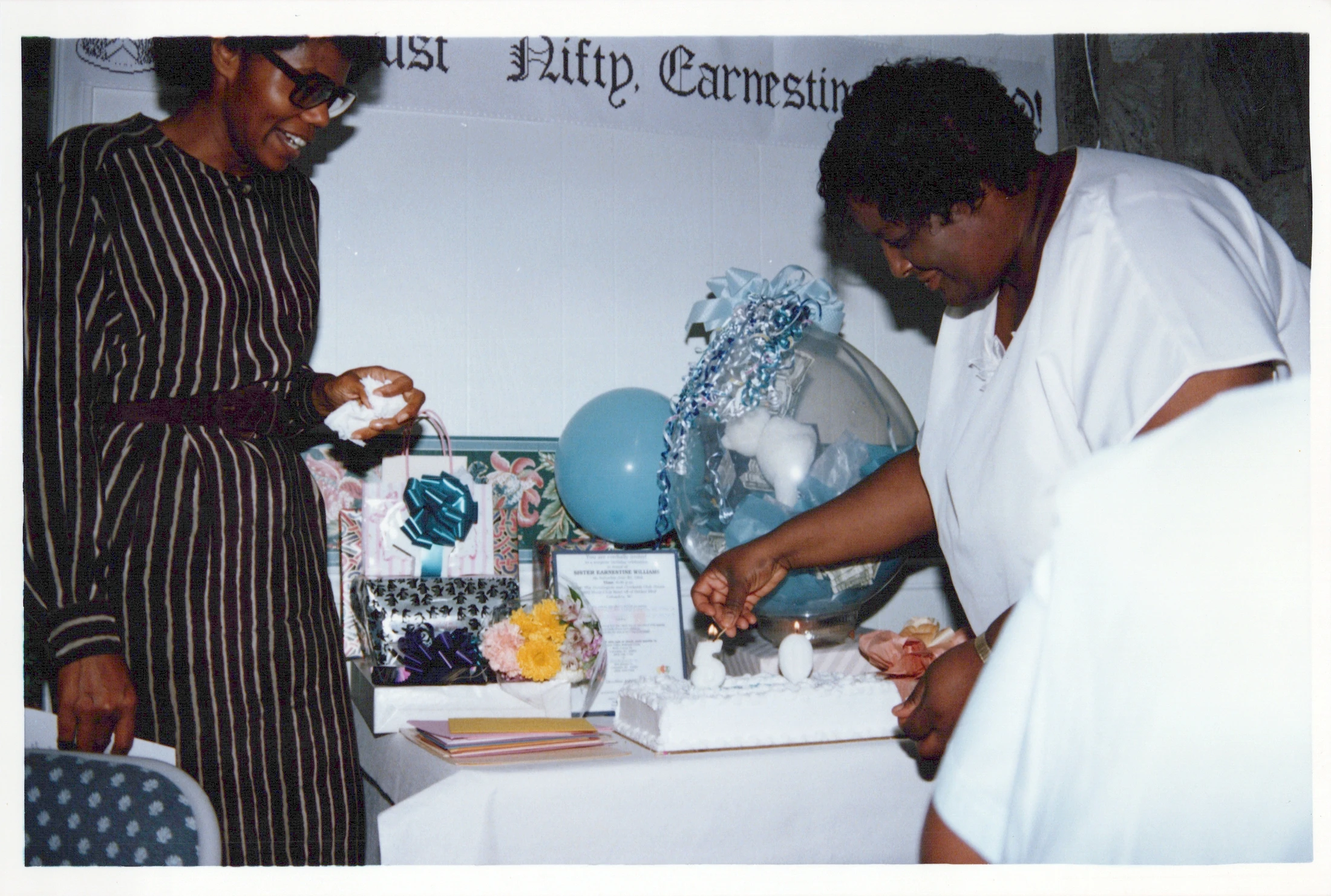 Two Black women at a party. One lights candles on a white cake while the other smiles. A banner, gifts, and balloons decorate the celebratory table.