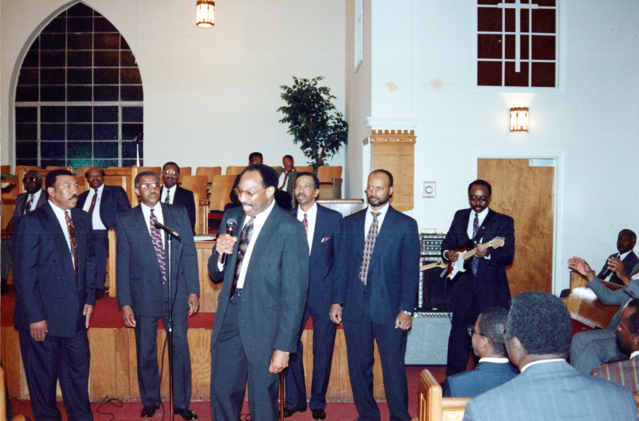 A group of Black men in dark suits perform in a church. One man sings into a microphone, while others sing behind him and one plays an electric guitar.