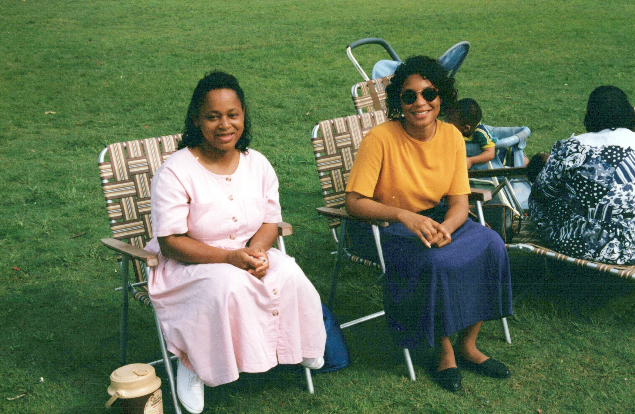 A vintage photograph of two women sitting in folding chairs on a green lawn, smiling. One wears a pink dress, the other a yellow shirt and sunglasses.