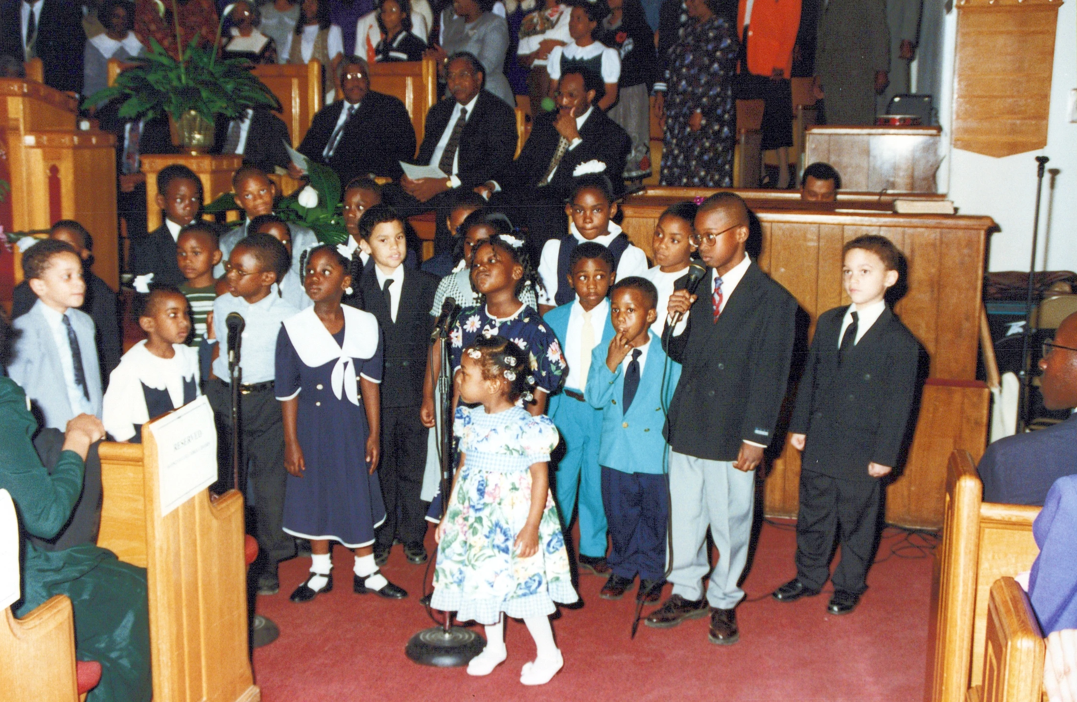 A large group of young children dressed in their Sunday best stand at the front of a church, performing for the congregation with microphones.