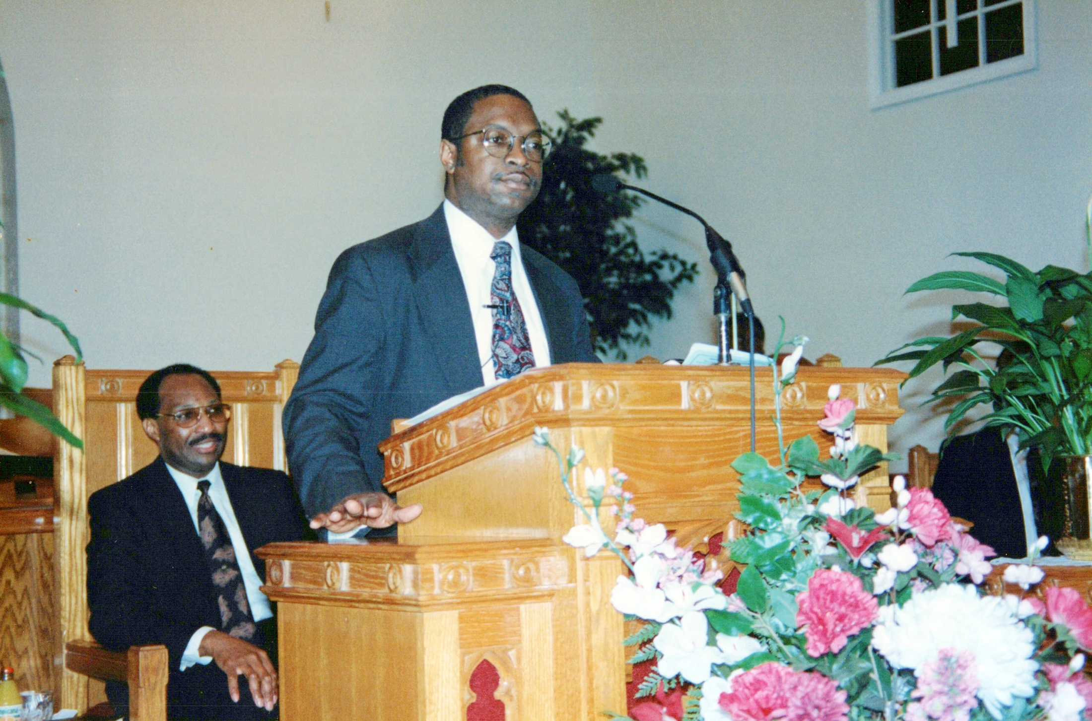 A Black man wearing glasses and a suit speaks at a wooden pulpit in a church. Another man in a suit sits smiling behind him.