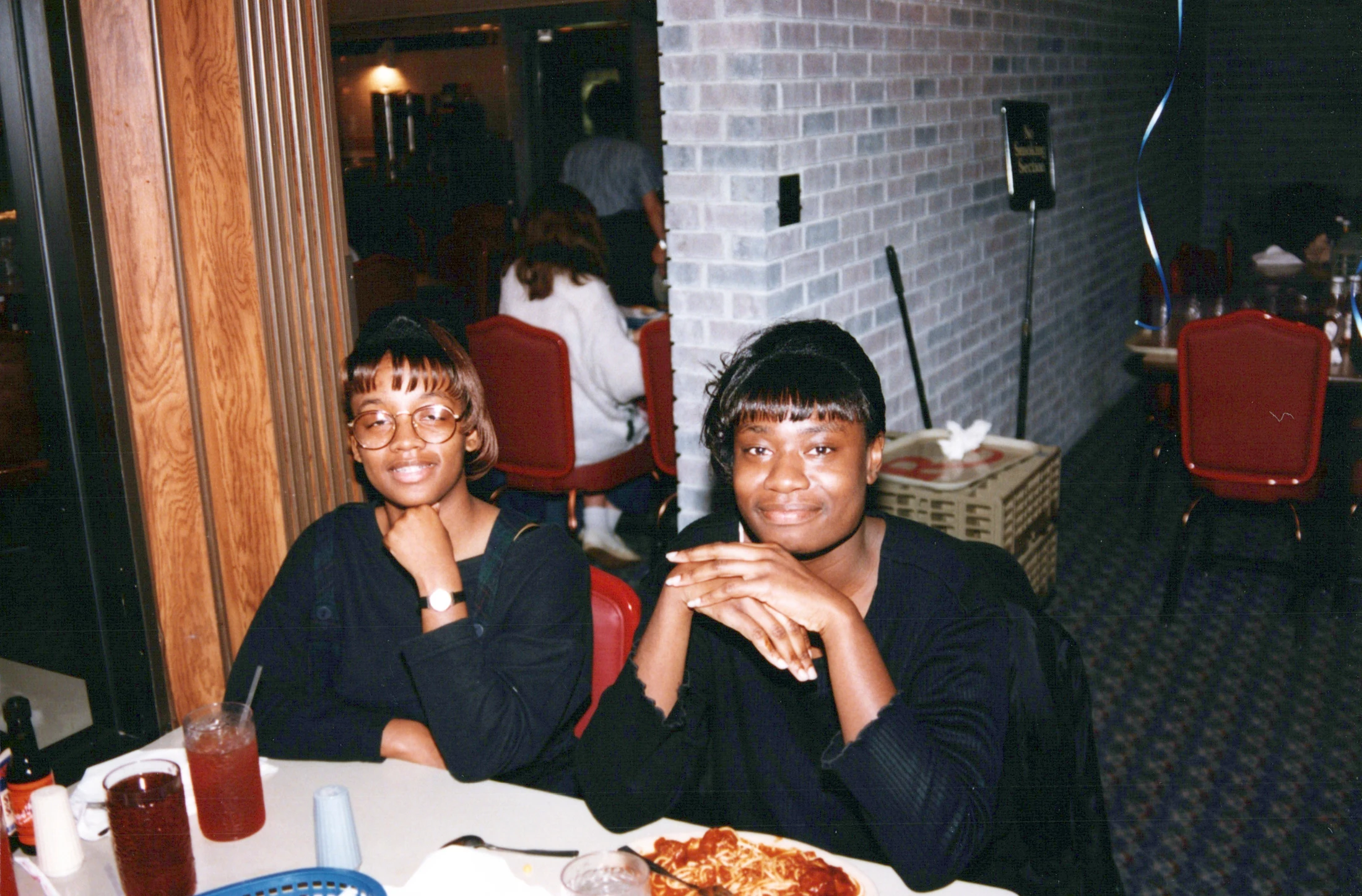 A vintage, slightly grainy photograph of two young Black women sitting at a dinner table in a restaurant, smiling for the camera.
