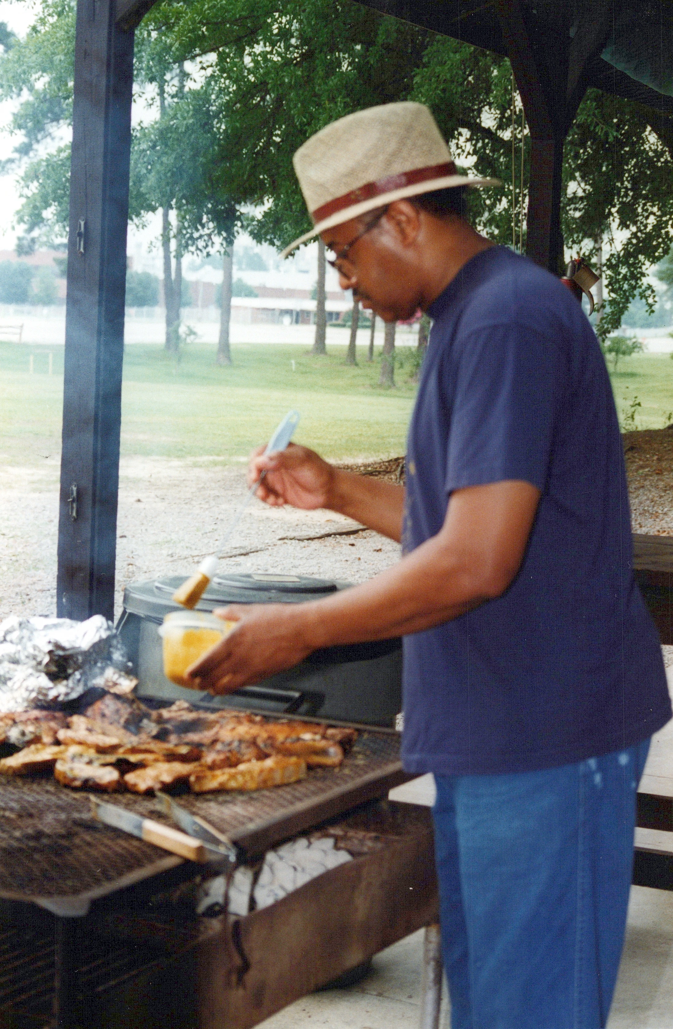 A man wearing a fedora and glasses stands at a charcoal grill, brushing a yellow sauce onto pieces of meat as they cook outdoors under a pavilion.